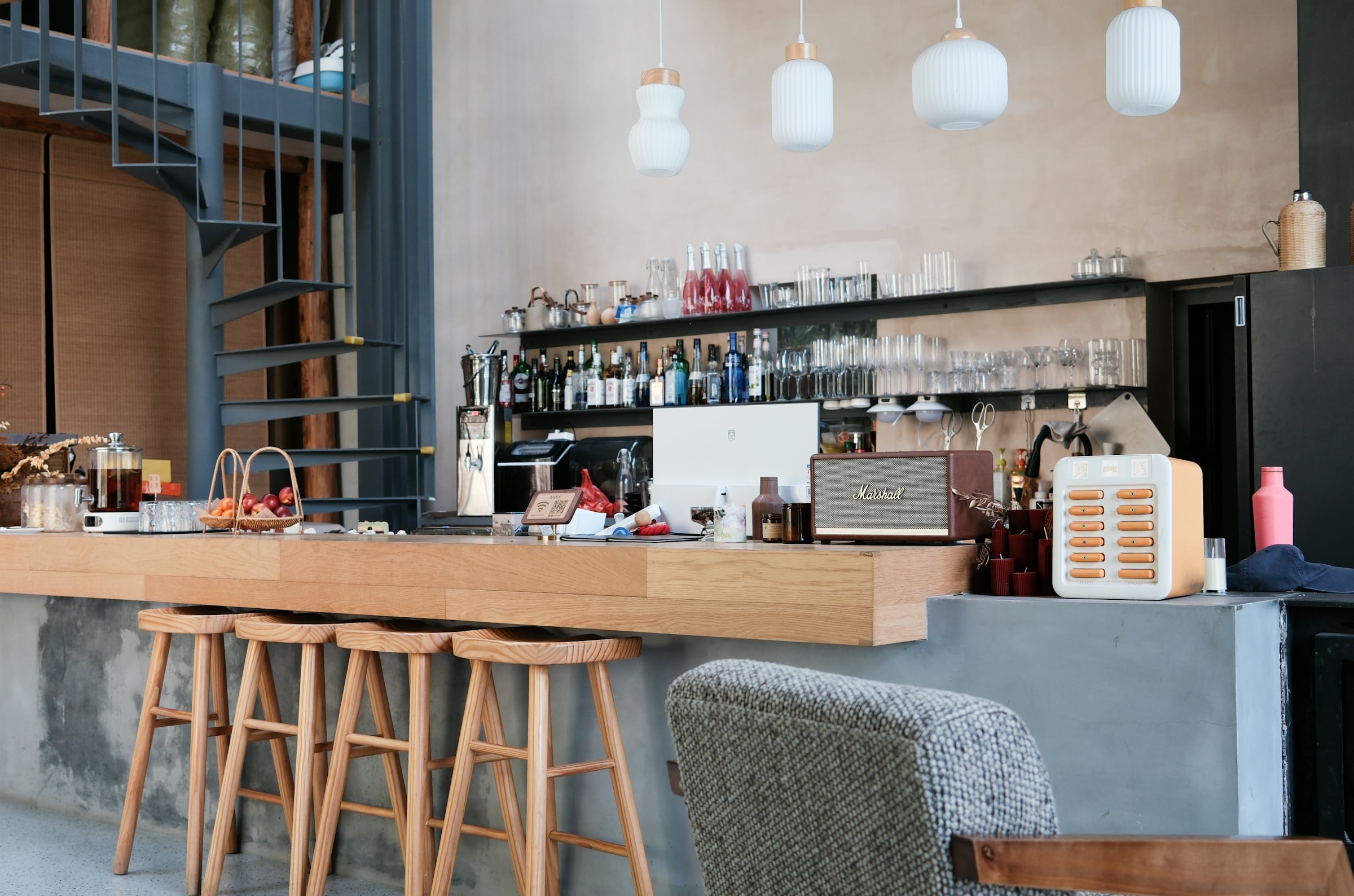 A modern loft-style bar with an open shelf of liquor bottles, hanging pendant lights, a spiral staircase and wooden bar stools along a concrete counter.