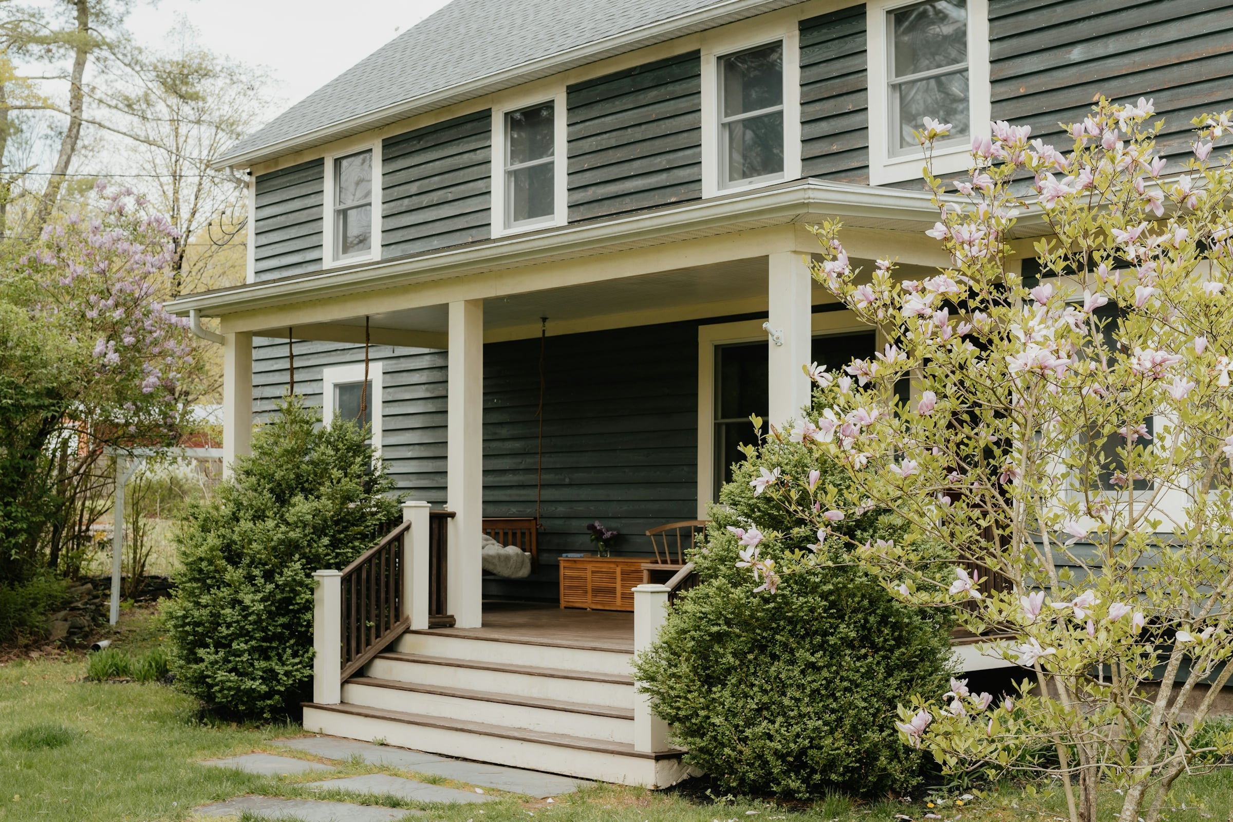 A pale green clapboard house with a covered front porch, white steps and a flowering magnolia beside it.