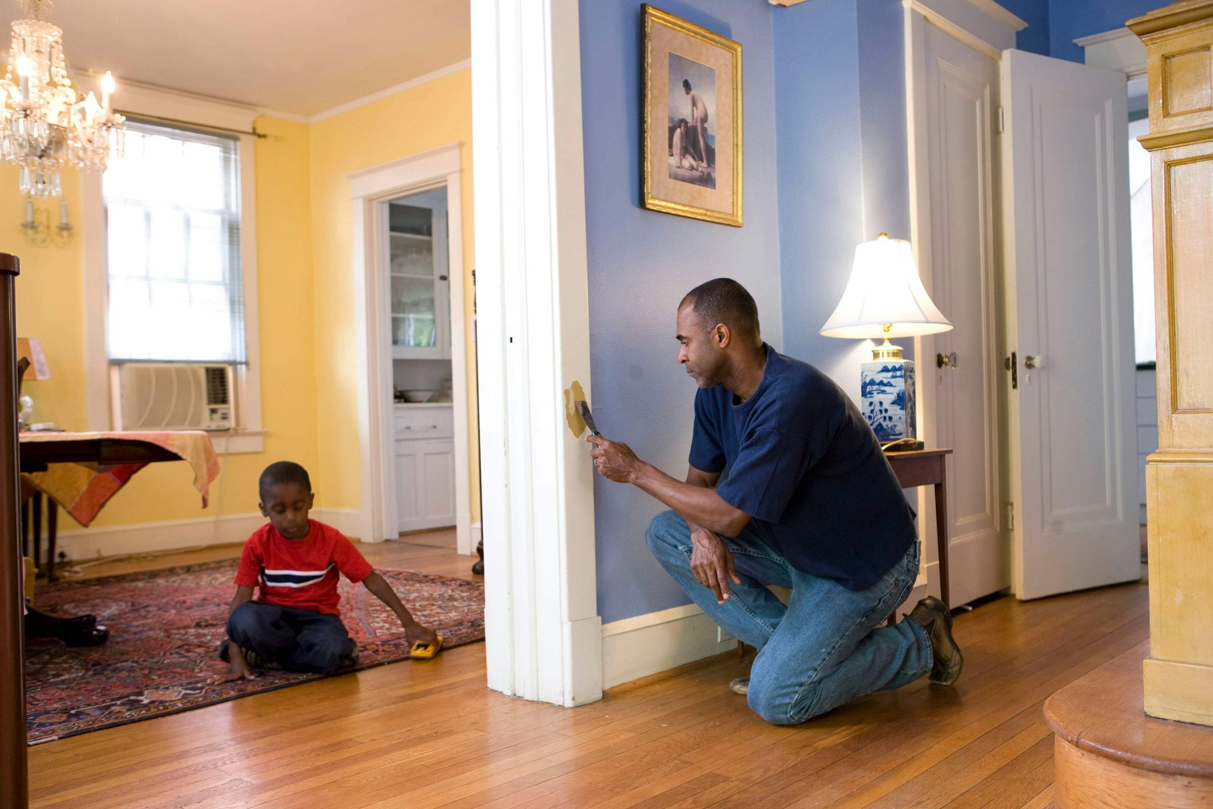 Man kneeling to examine a wall and baseboard inside a sunlit older home