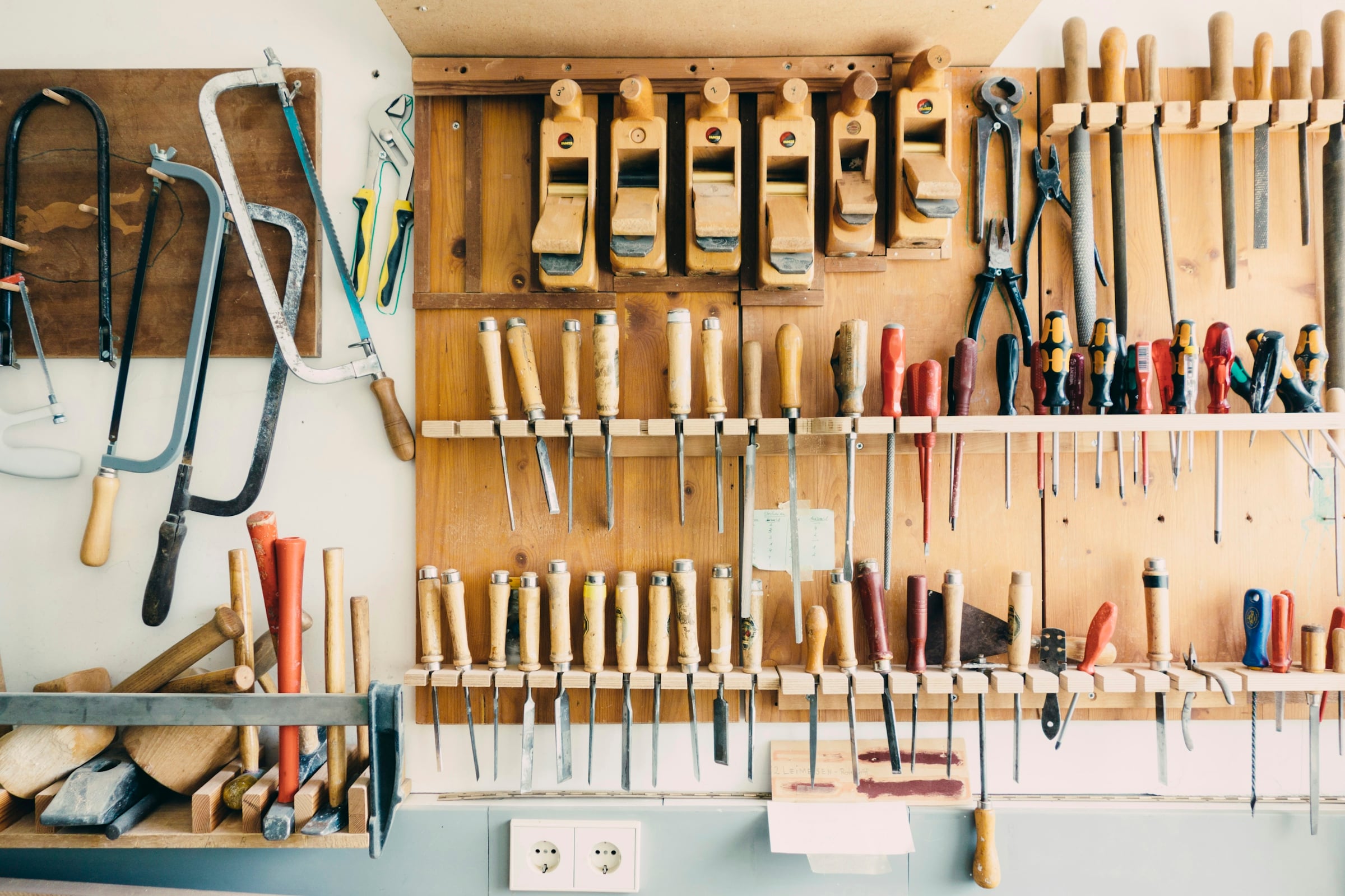 Hand tools including saws, clamps and chisels hung in rows on a workshop wall above a bench