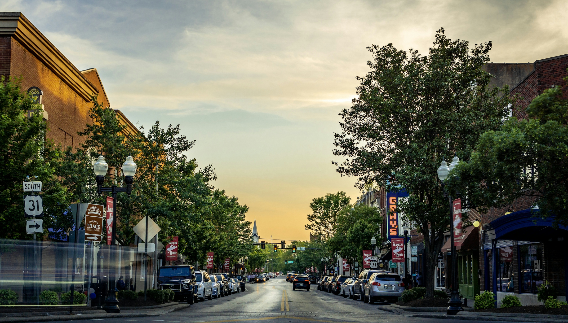 A small town main street at dusk, lined with parked cars, storefront signs and trees.