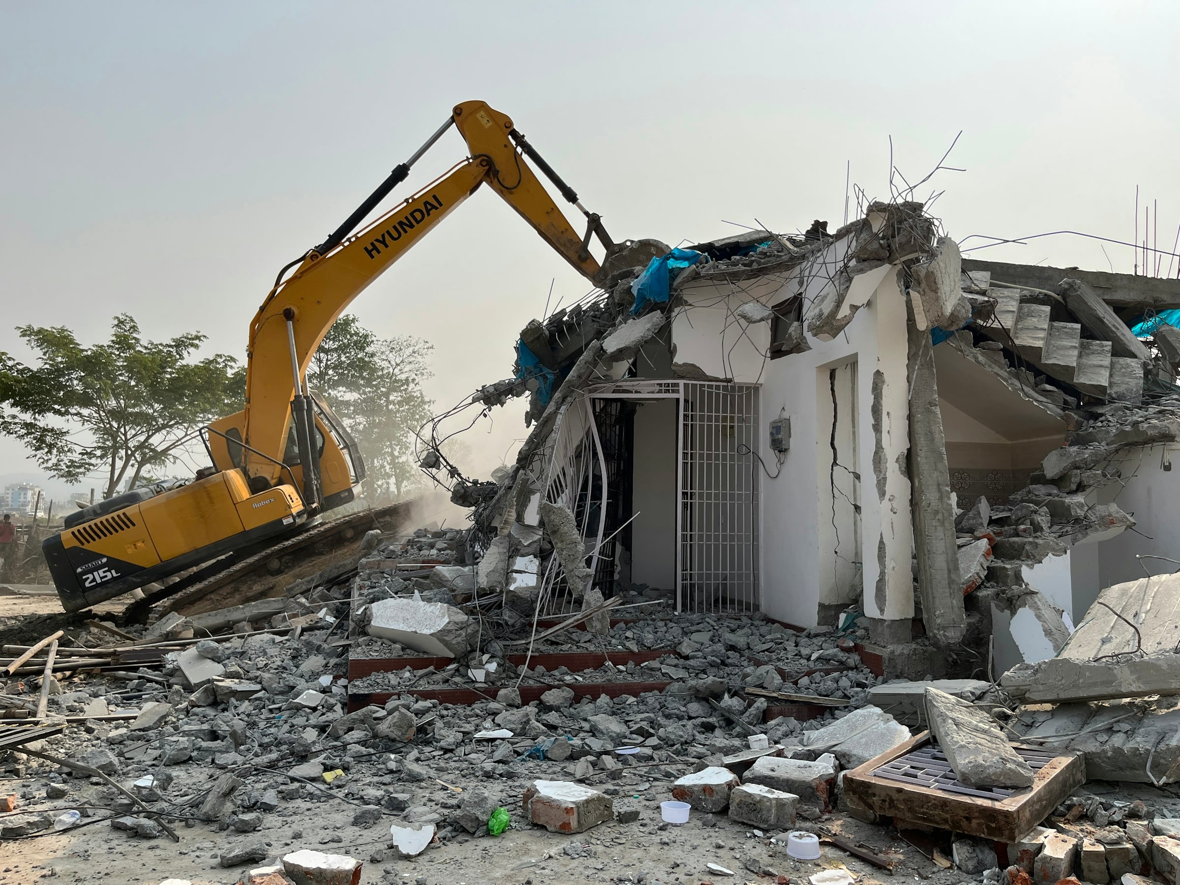 A yellow tracked excavator pulling down the upper storey of a damaged concrete building