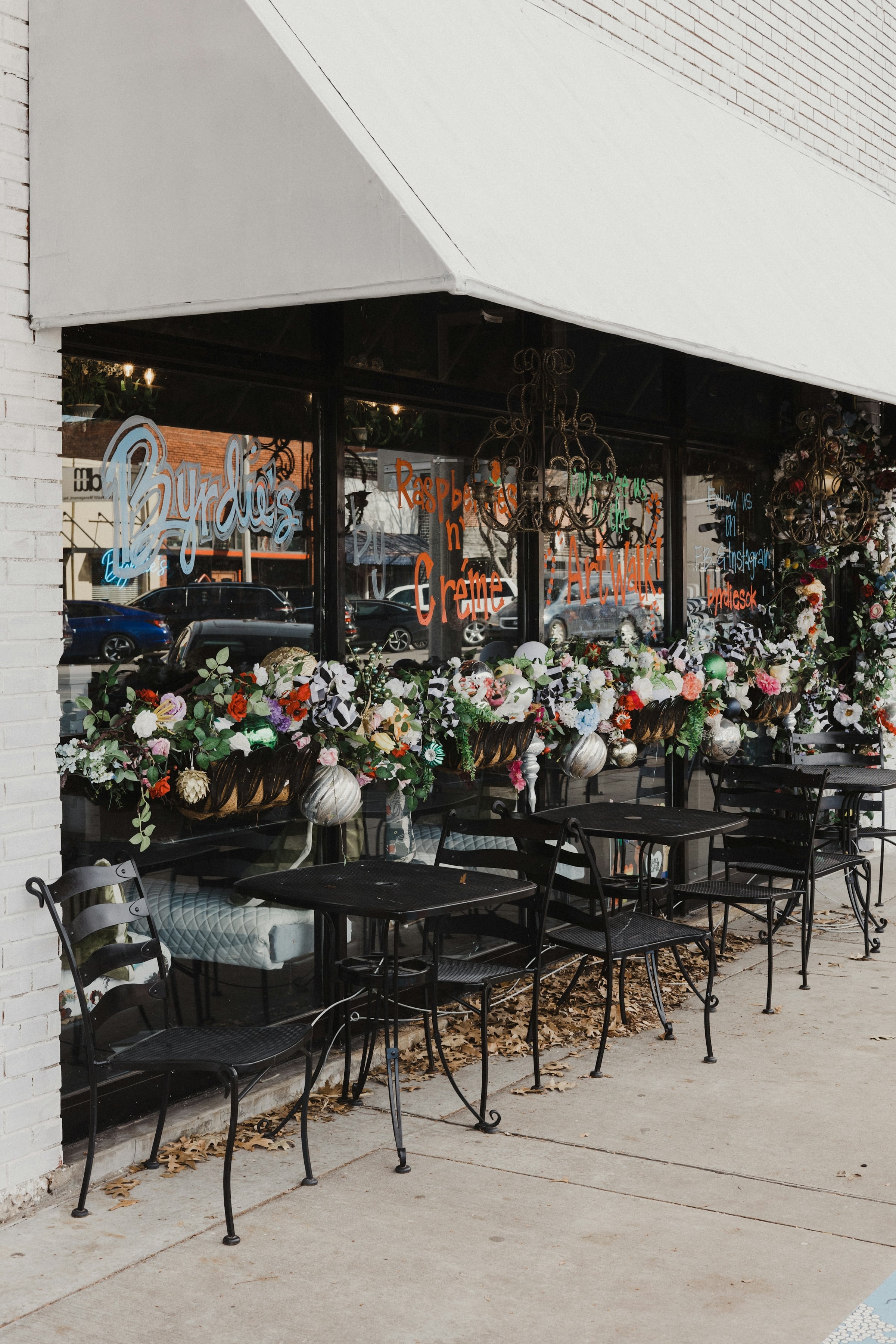 Storefront with a striped awning and large display windows, with flower arrangements lined up beside outdoor cafe tables