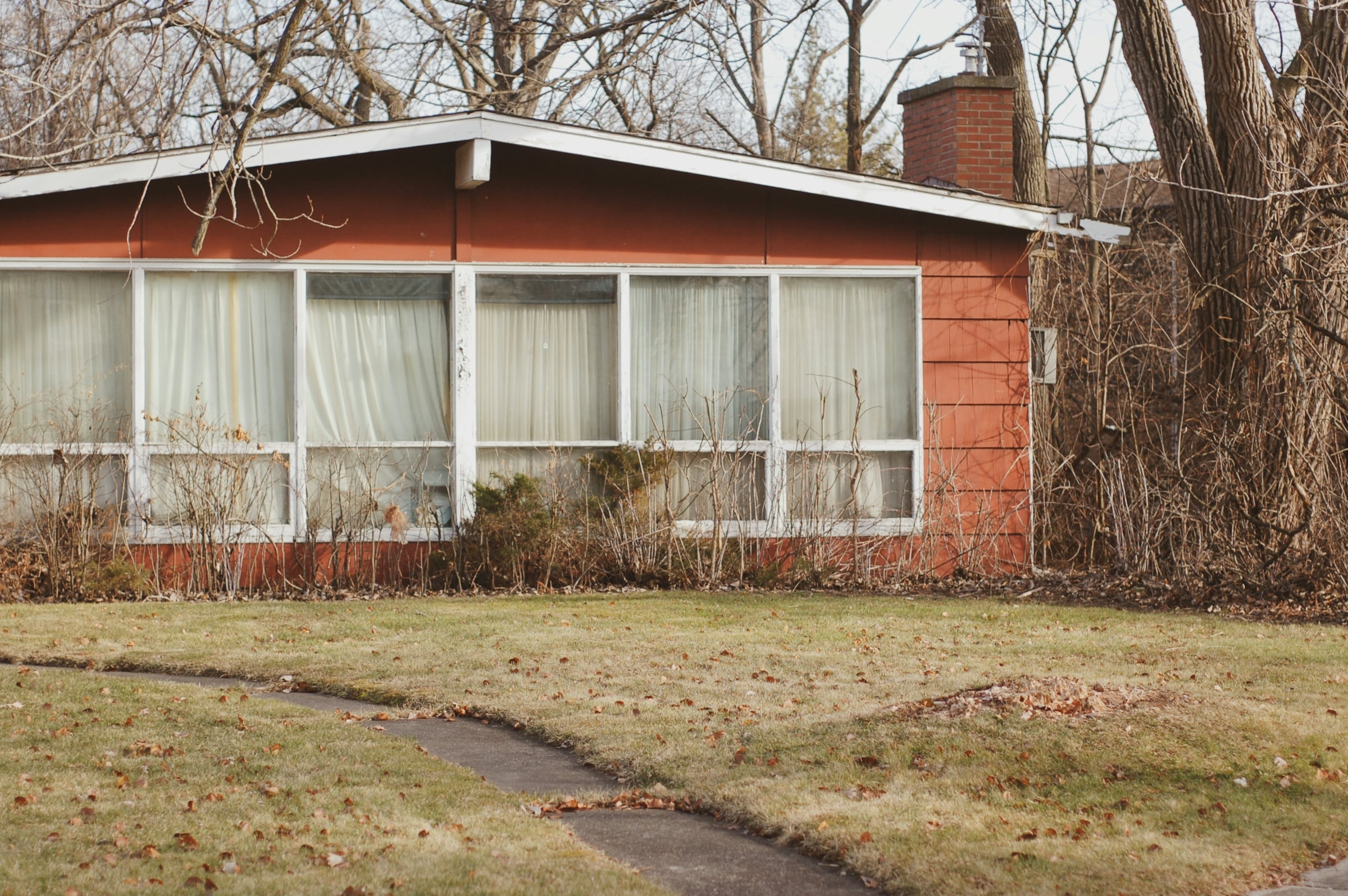 Low mid-century ranch house with a long band of windows, brick chimney and bare winter trees