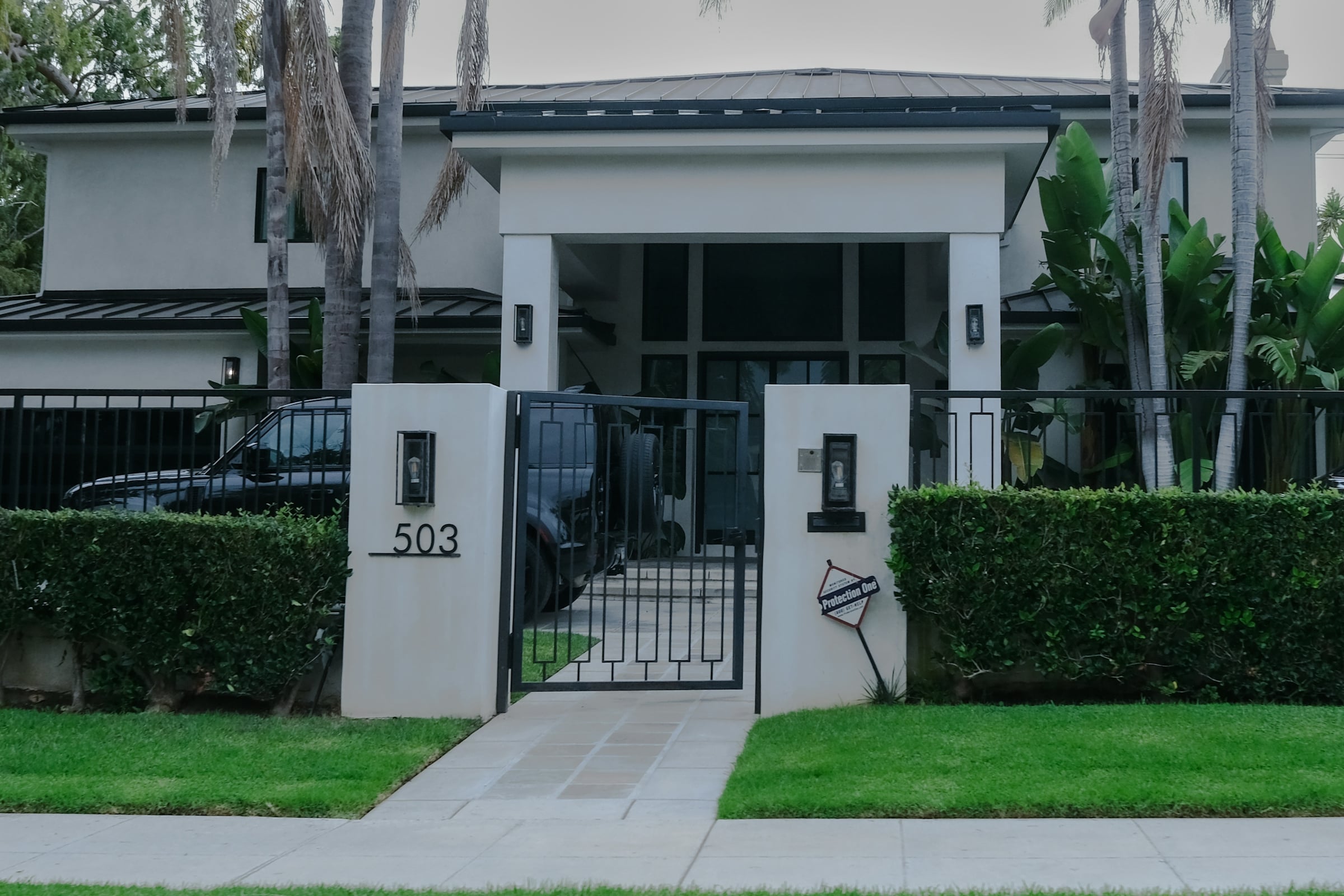 A flat roofed modern white house behind a clipped hedge and a metal driveway gate
