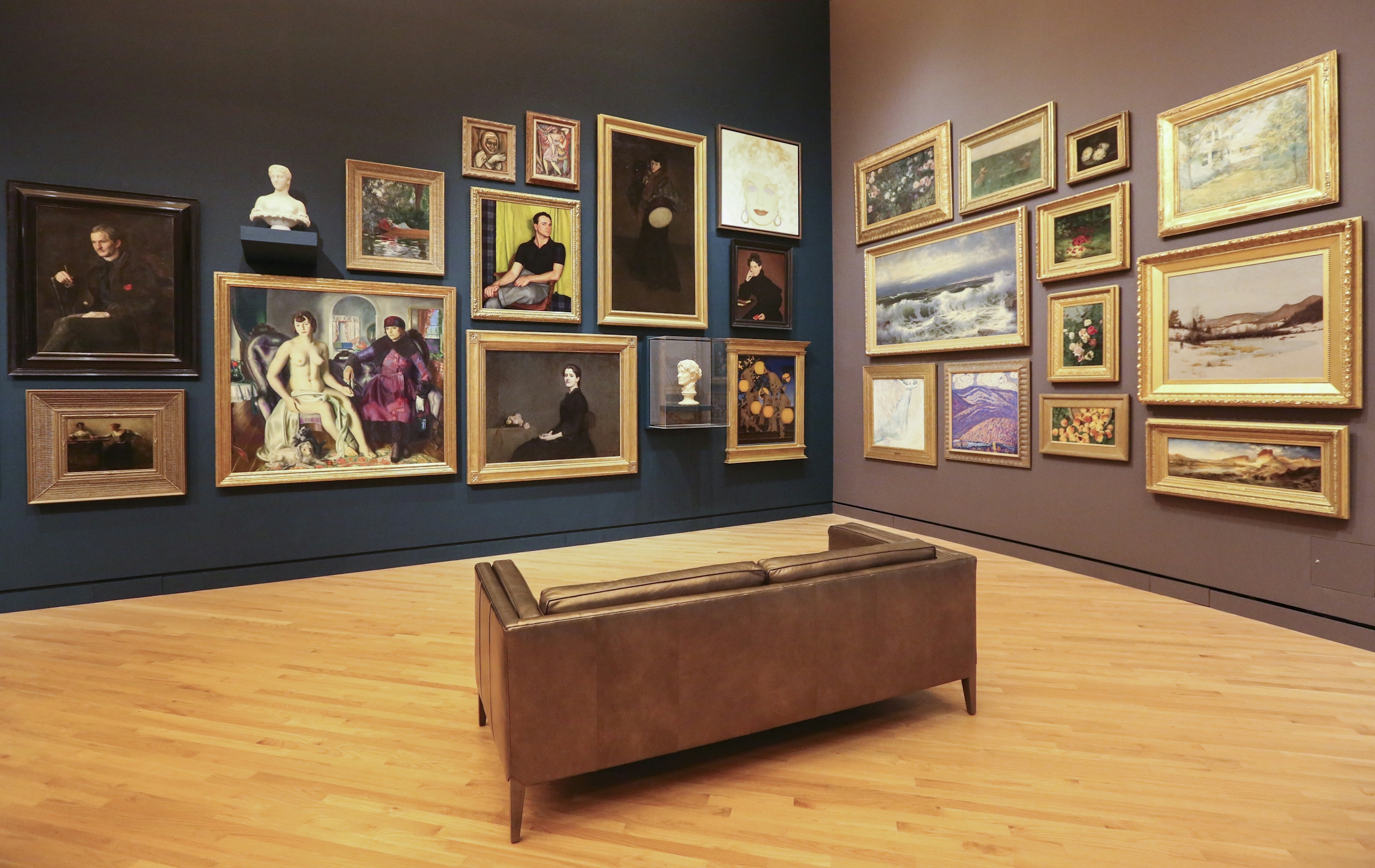 A museum gallery with paintings in gilt frames hung salon-style on a deep blue wall.