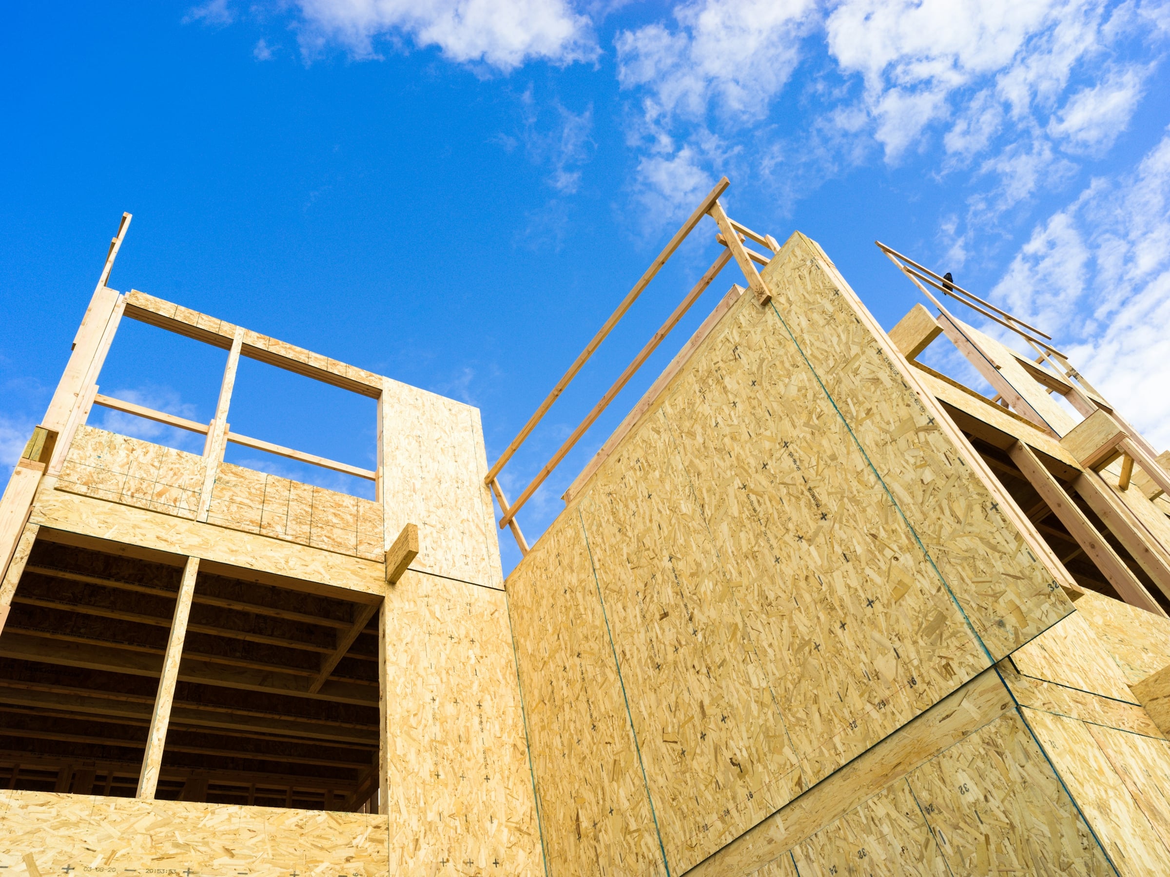A two-storey house under construction with exposed wood framing and OSB sheathing against a blue sky