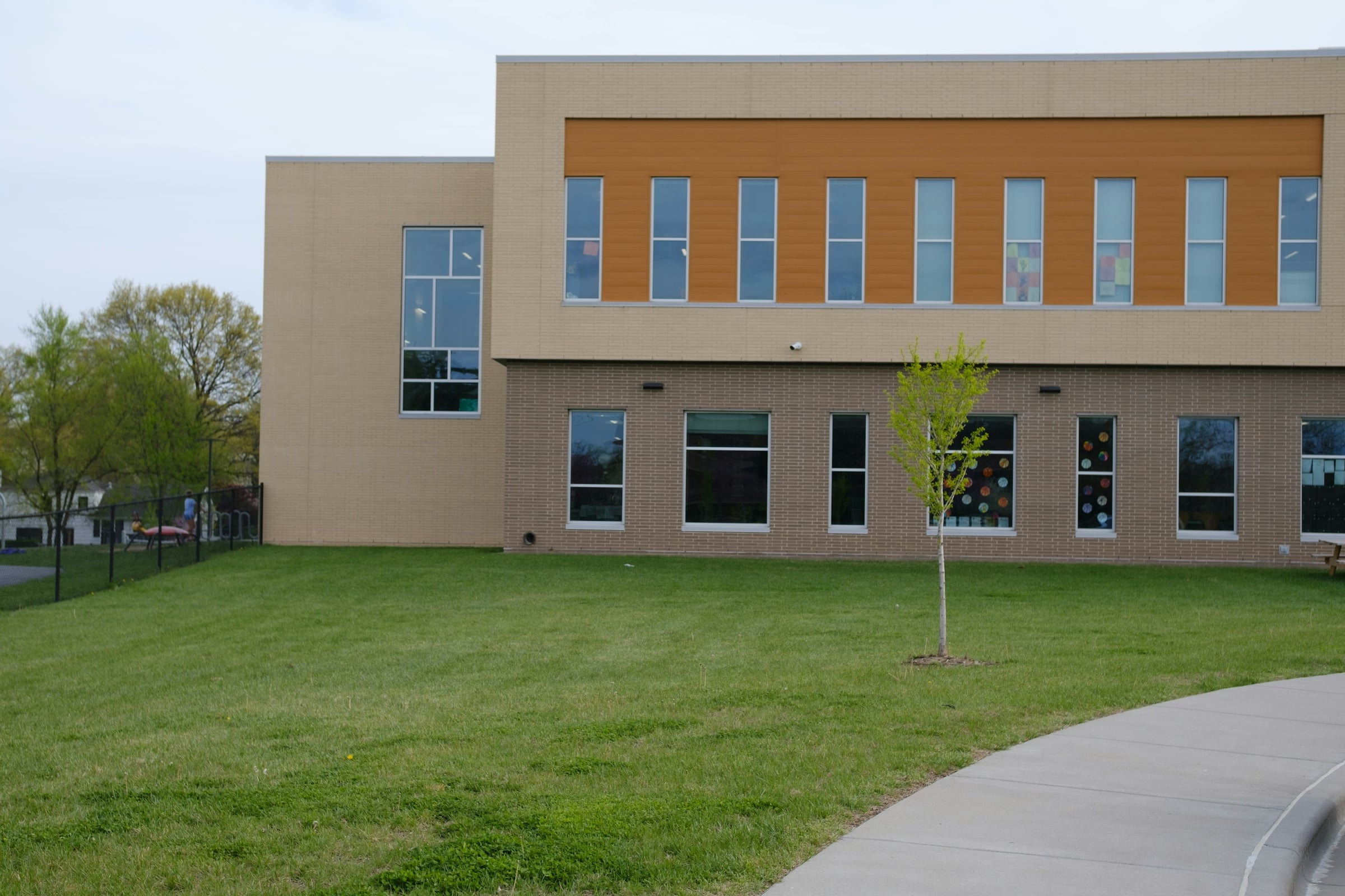 A modern brick elementary school building with an orange accent panel above a row of tall classroom windows, children's artwork visible inside, and a young tree planted on the lawn out front.