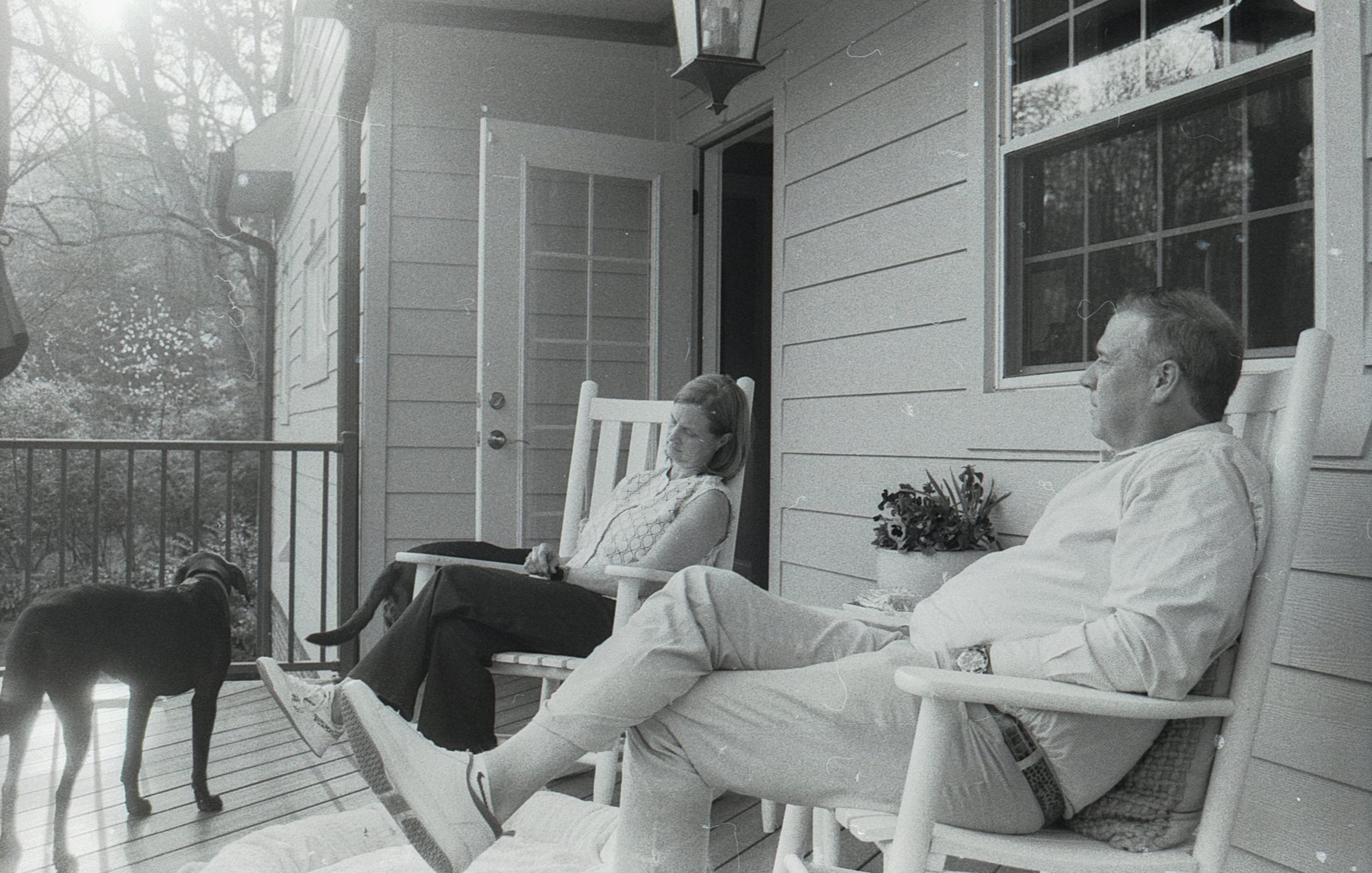 An older couple sitting in rocking chairs on a covered front porch with a dog lying on the boards beside them