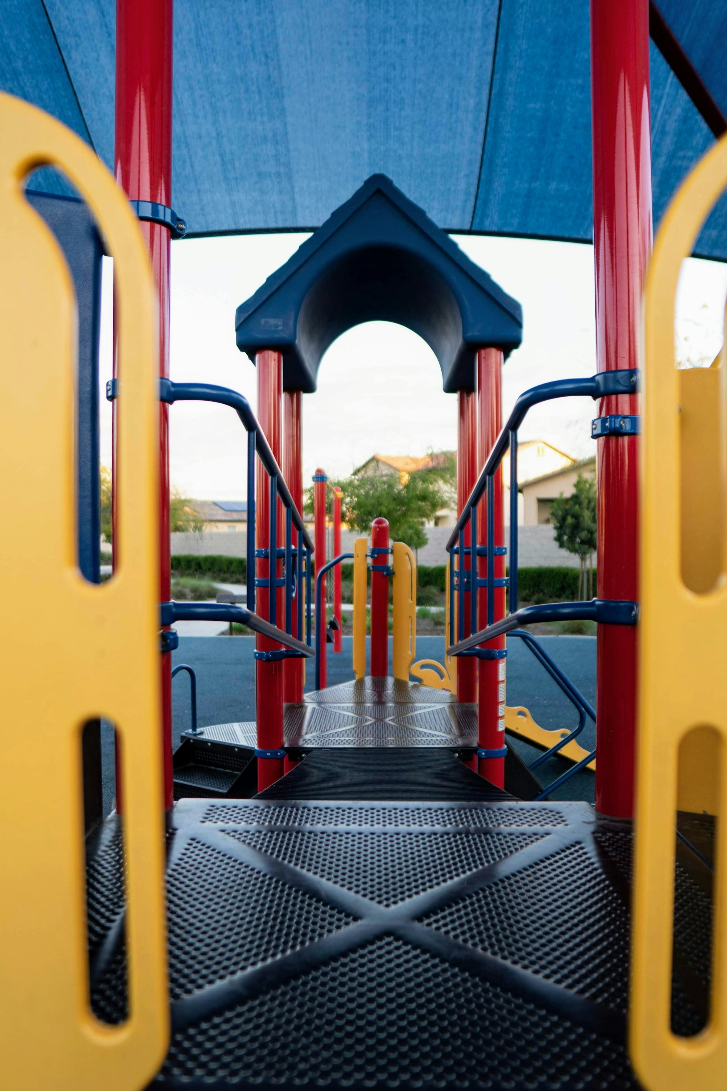 A children's playground structure with yellow and blue panels and a metal slide, with houses and trees behind it.