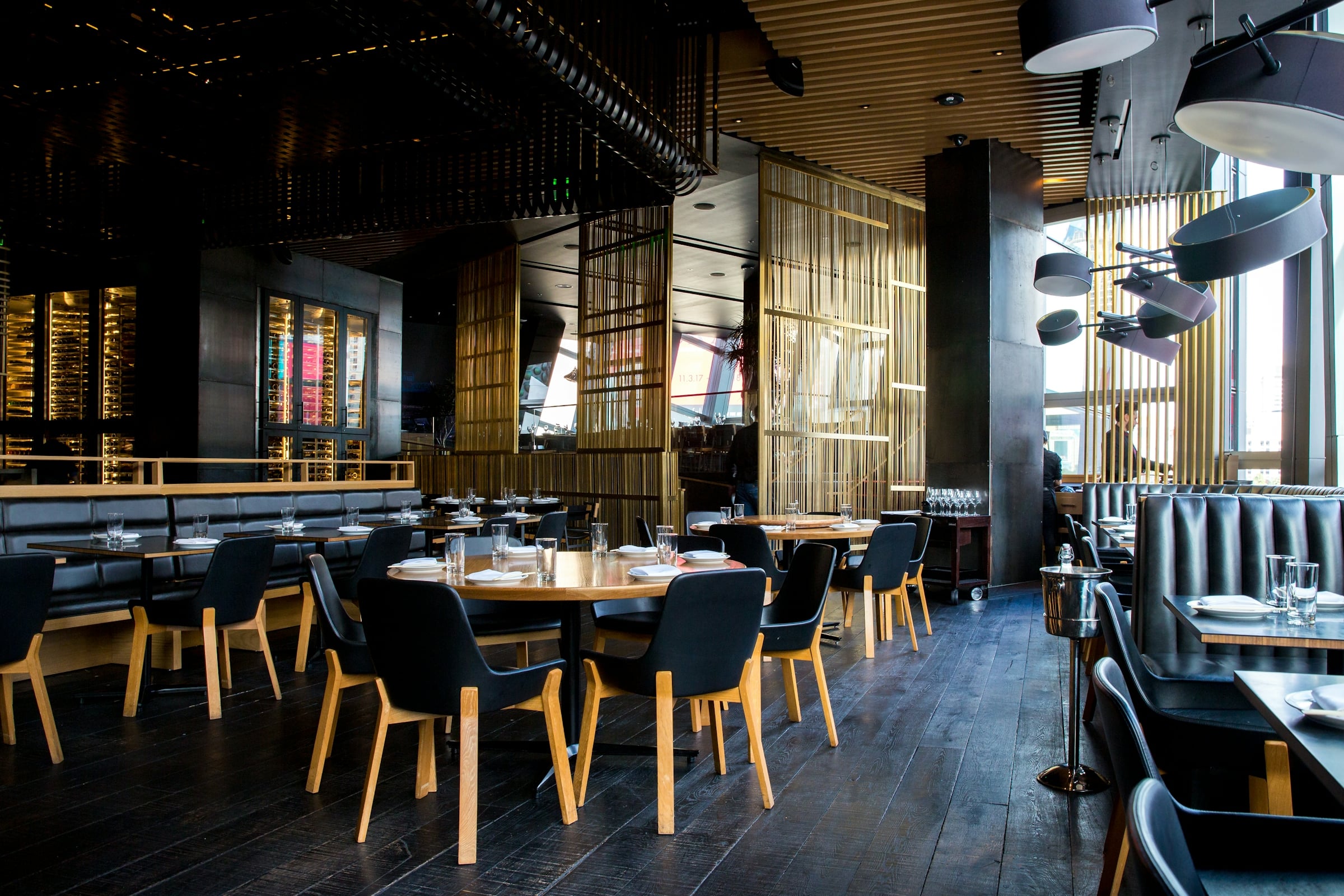 The interior of a modern restaurant with round wooden tables, dark upholstered chairs and pendant lighting.
