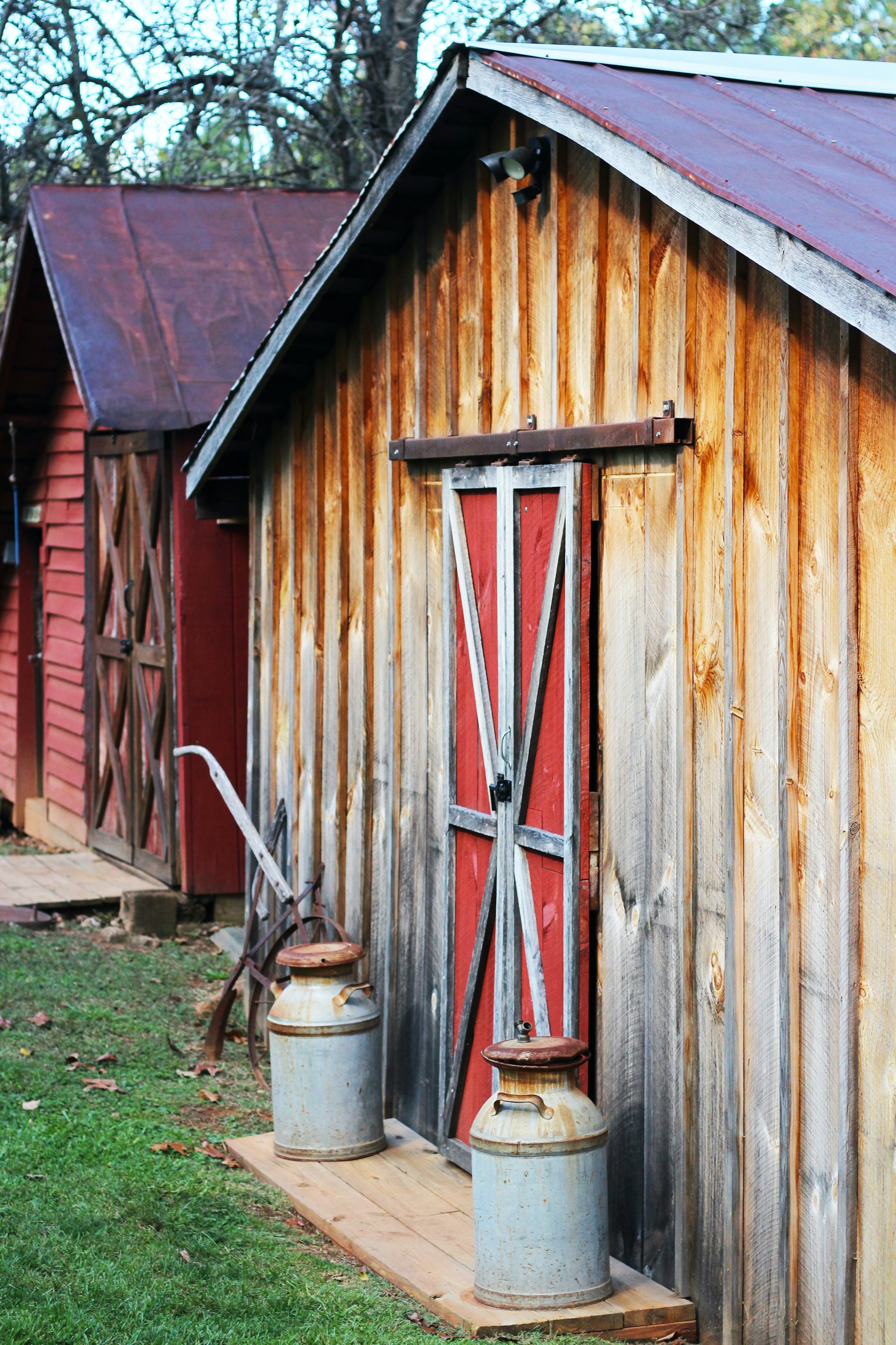 A weathered wooden outbuilding with a red barn door and a metal roof on a rural property, with two old metal milk cans set on the porch.