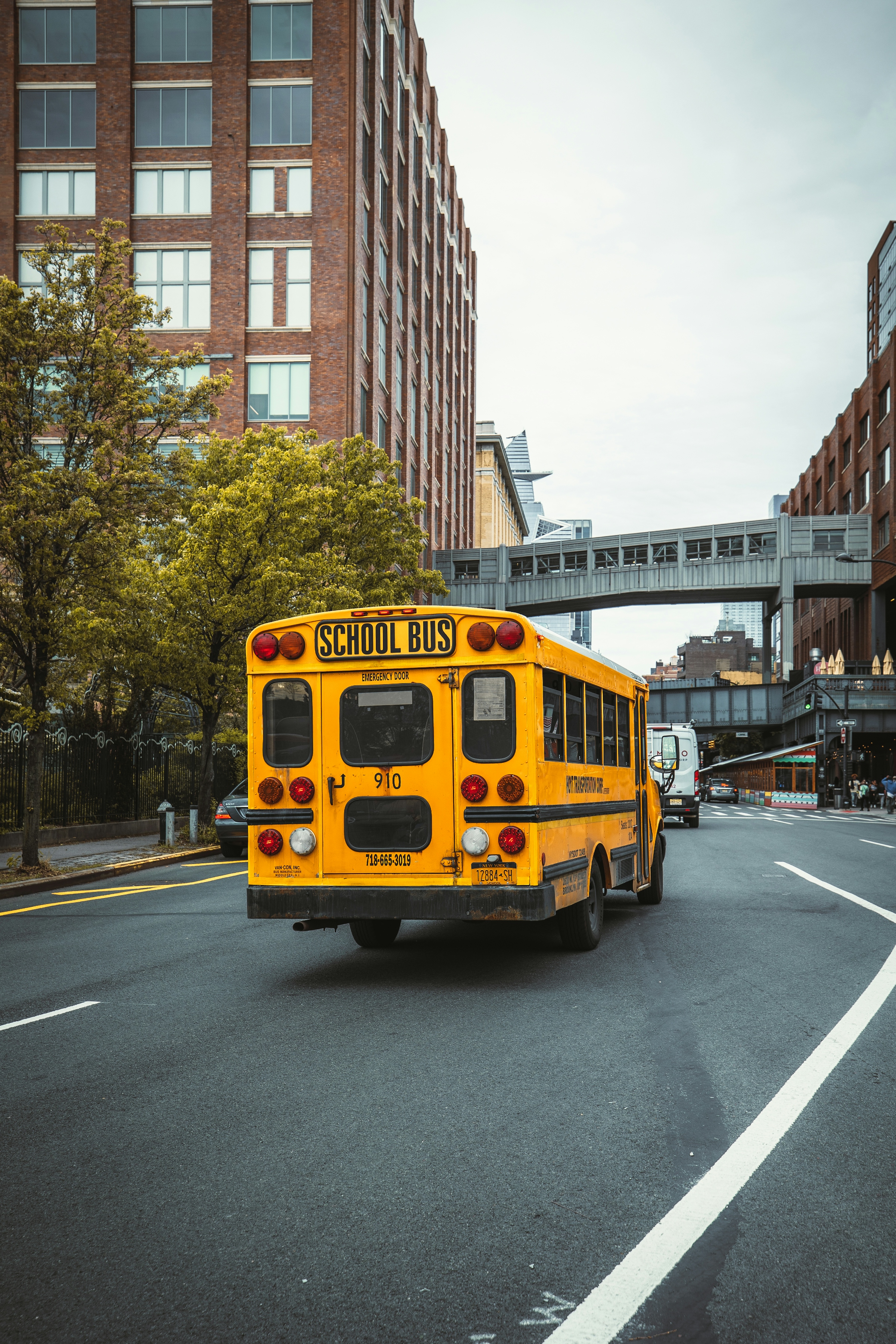 The back of a yellow school bus on a city street, with brick buildings and an overhead walkway.