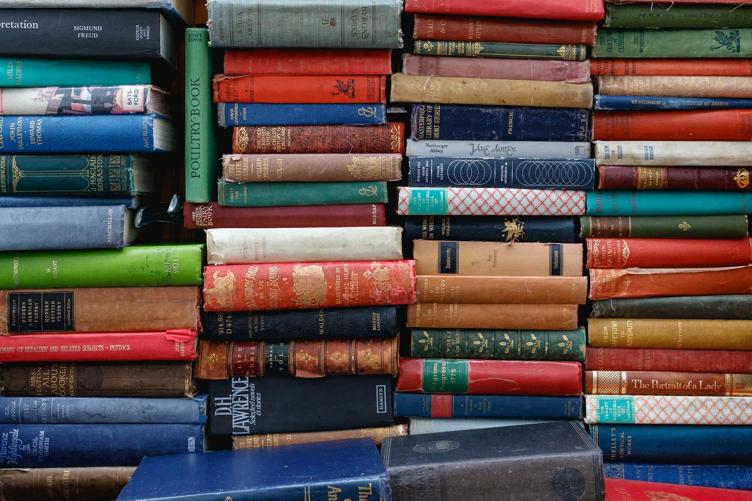 A tall stack of hardback books with worn cloth spines in red, blue and green.