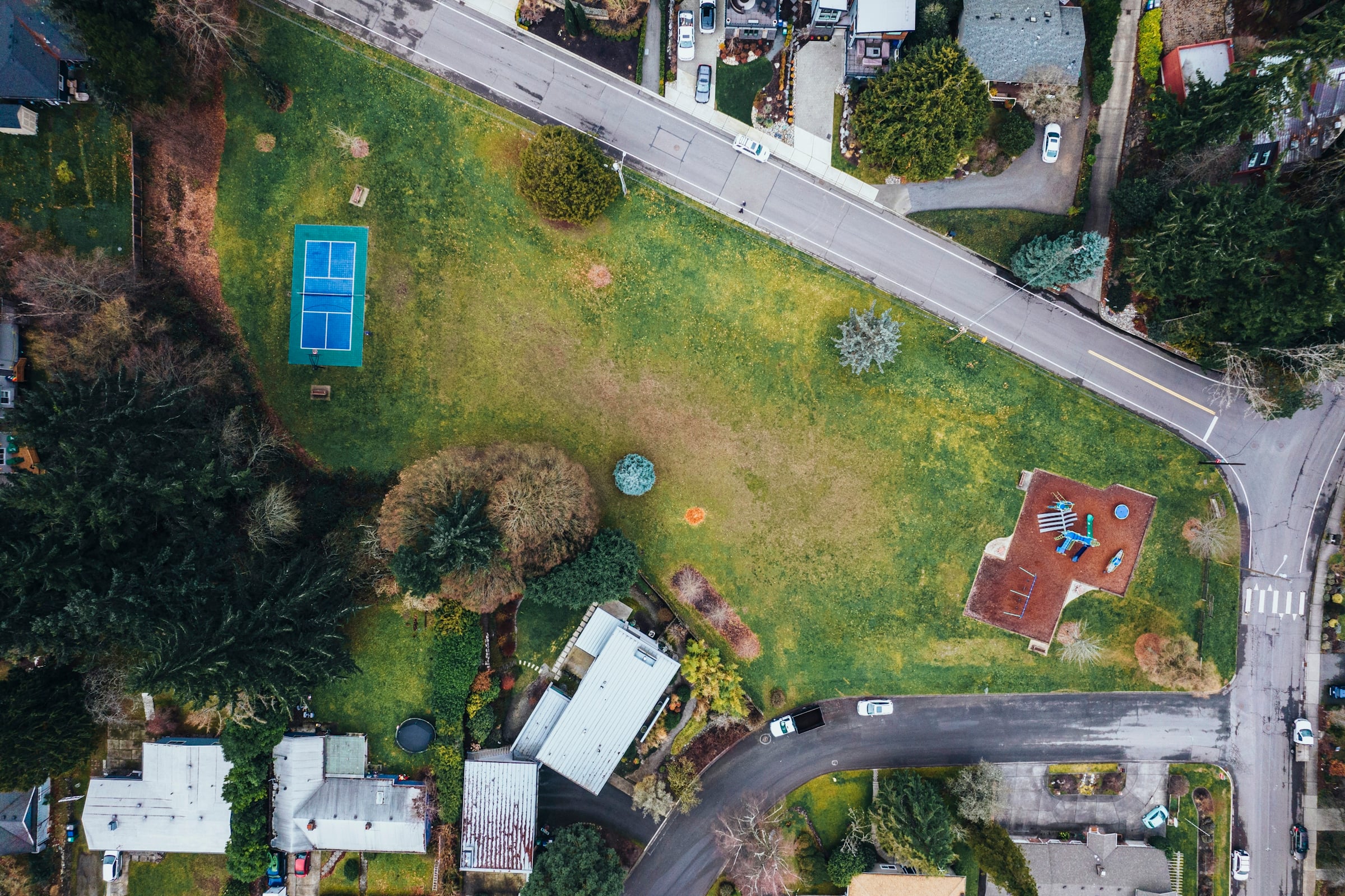 An overhead view of a suburban neighborhood showing houses, mature trees, a winding street and a blue tennis court.