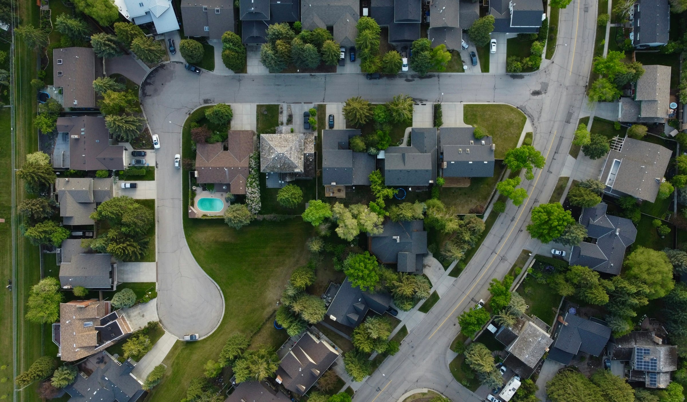 Overhead view of a suburban cul-de-sac lined with single-family houses, paved driveways and mature leafy trees between back yards