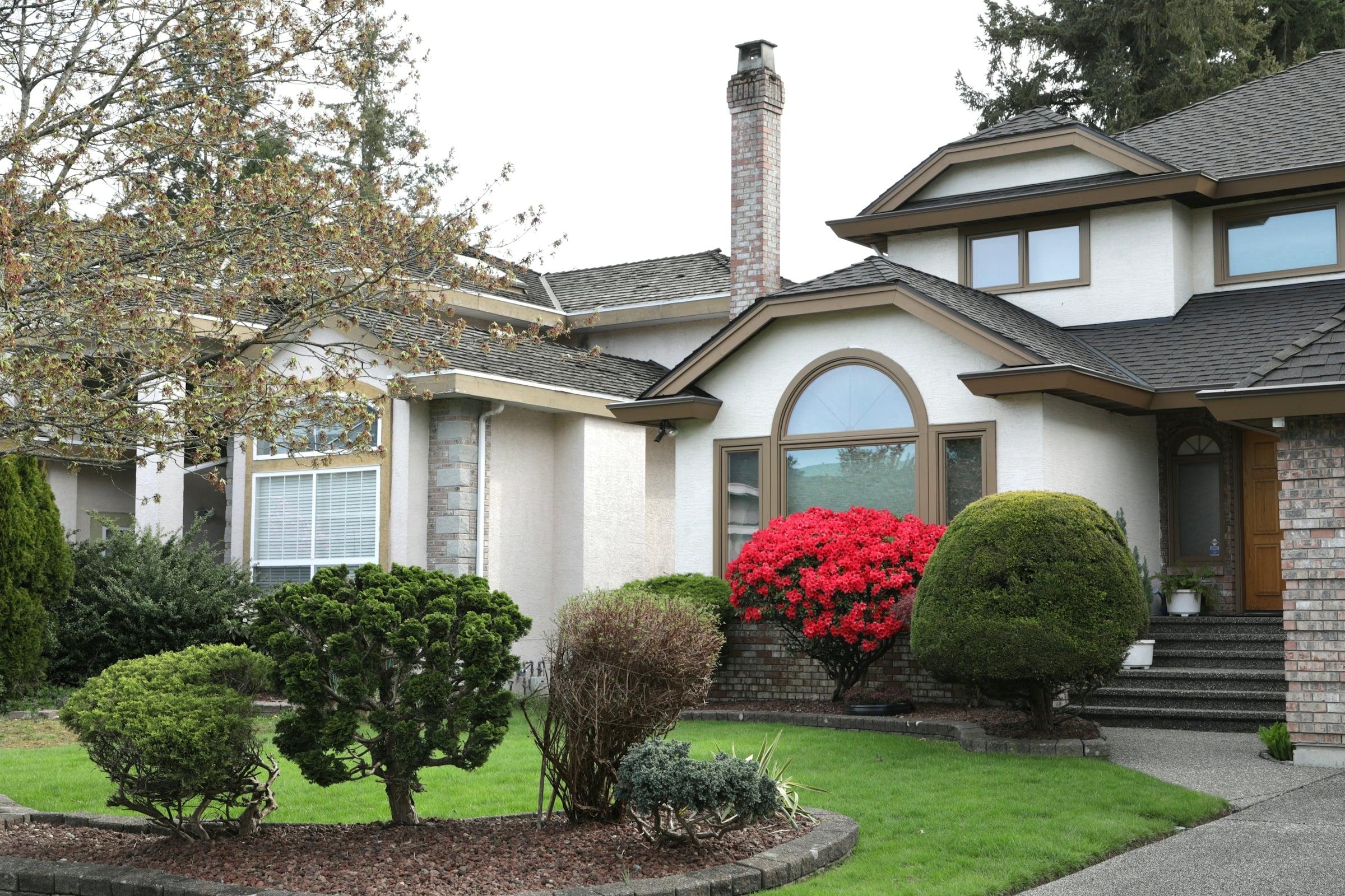 A large suburban house with a stone and stucco front, an arched window, clipped boxwood and flowering azaleas along the front walk.