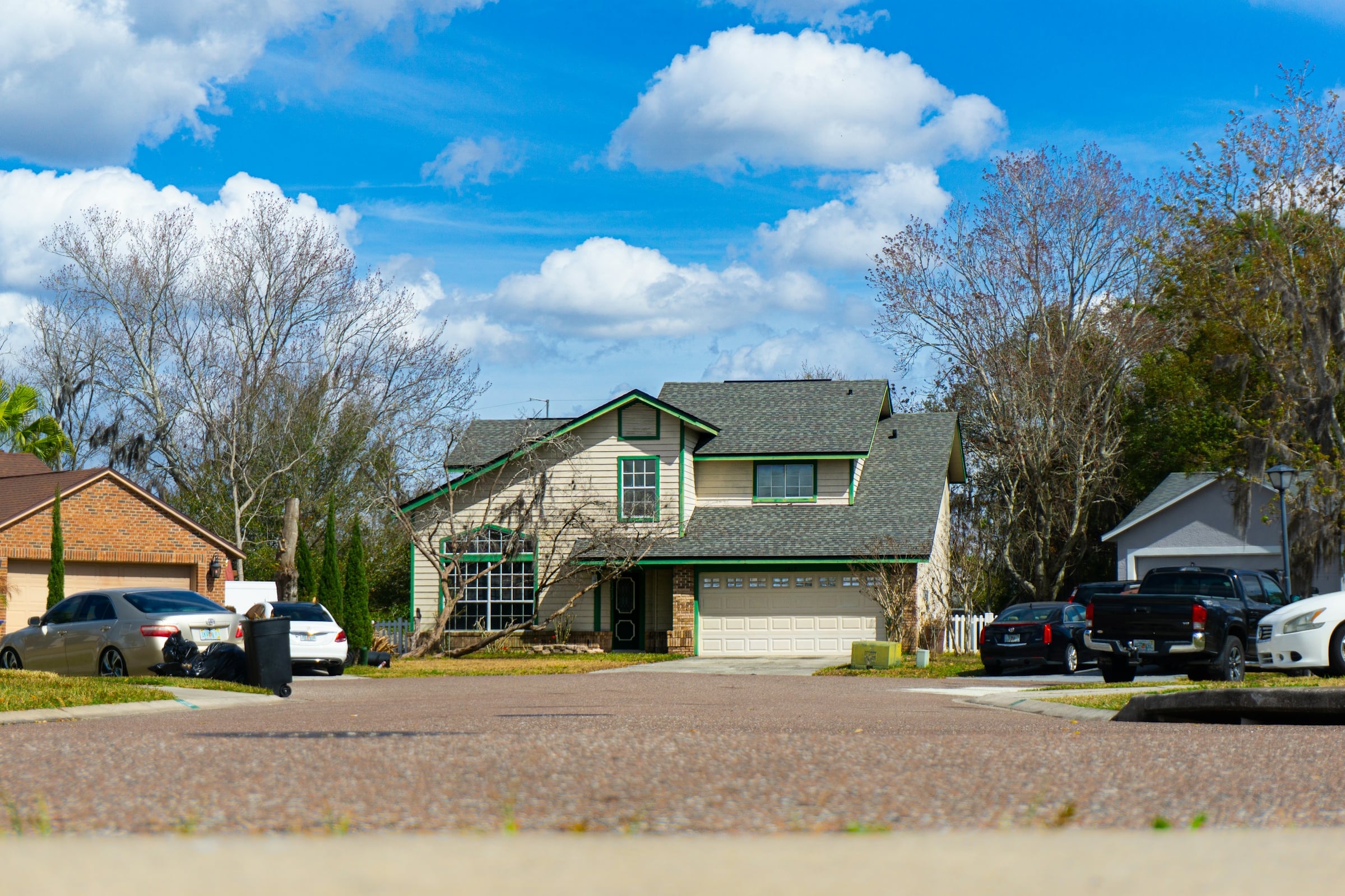A two-story suburban house with an attached garage and a wide driveway, with cars parked under a bright blue sky.