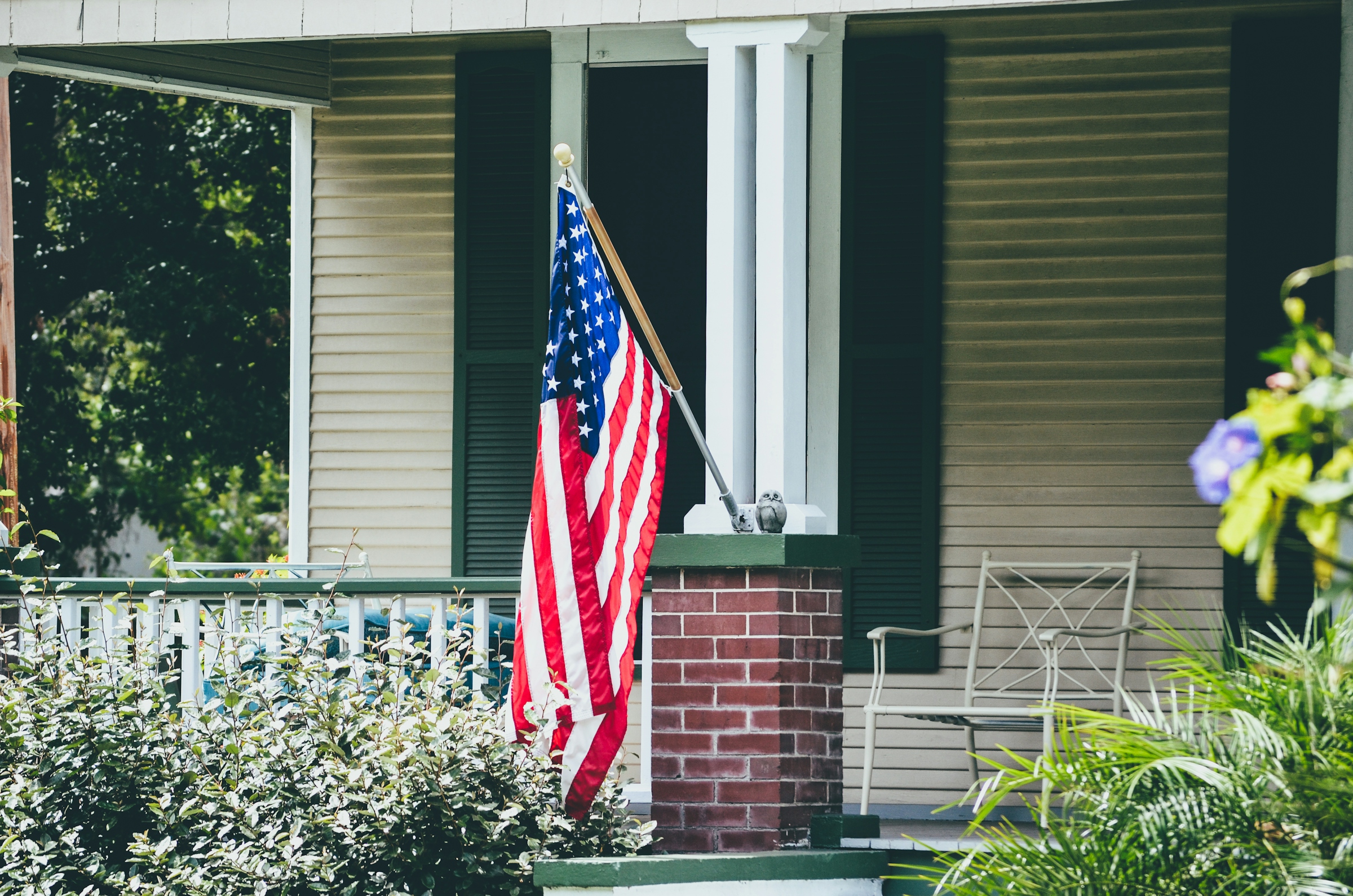 A United States flag mounted on a brick porch column beside a white porch rail and a wooden rocking chair.