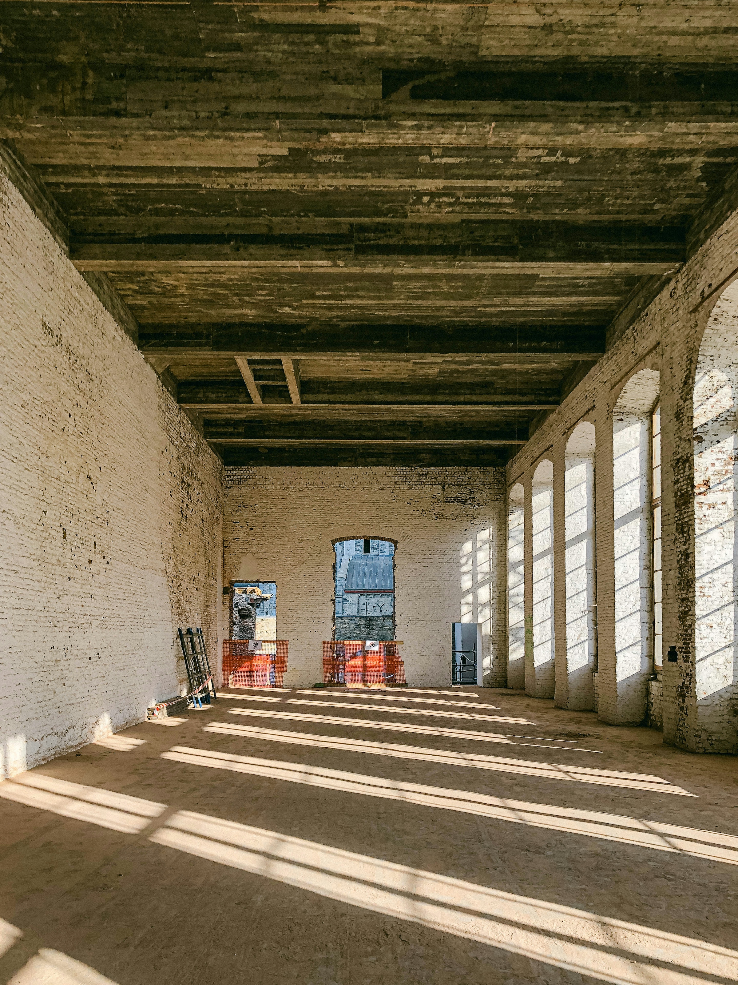 The exposed brick walls and heavy timber ceiling of an old industrial building mid-renovation, with arched windows letting in sunlight across a bare concrete floor.