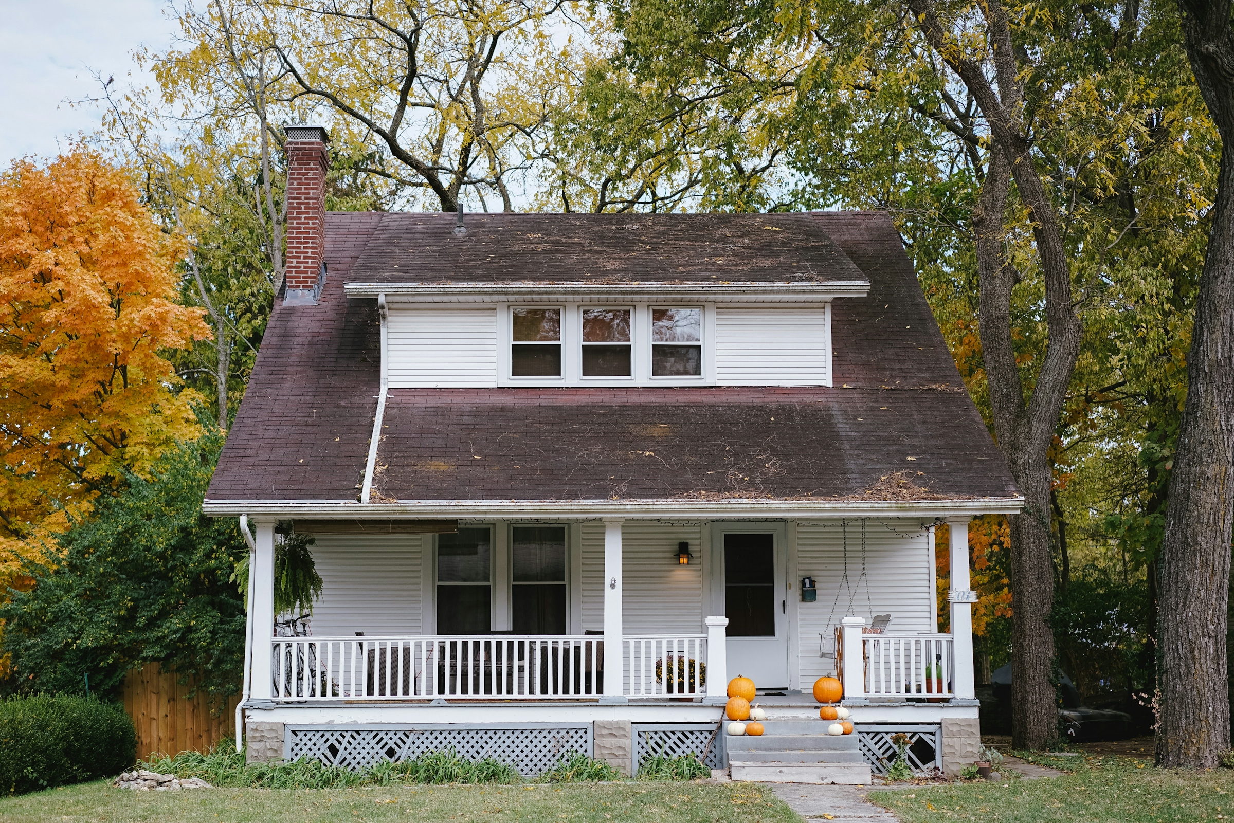 A white American bungalow with a deep front porch, tapered porch columns and pumpkins on the steps.