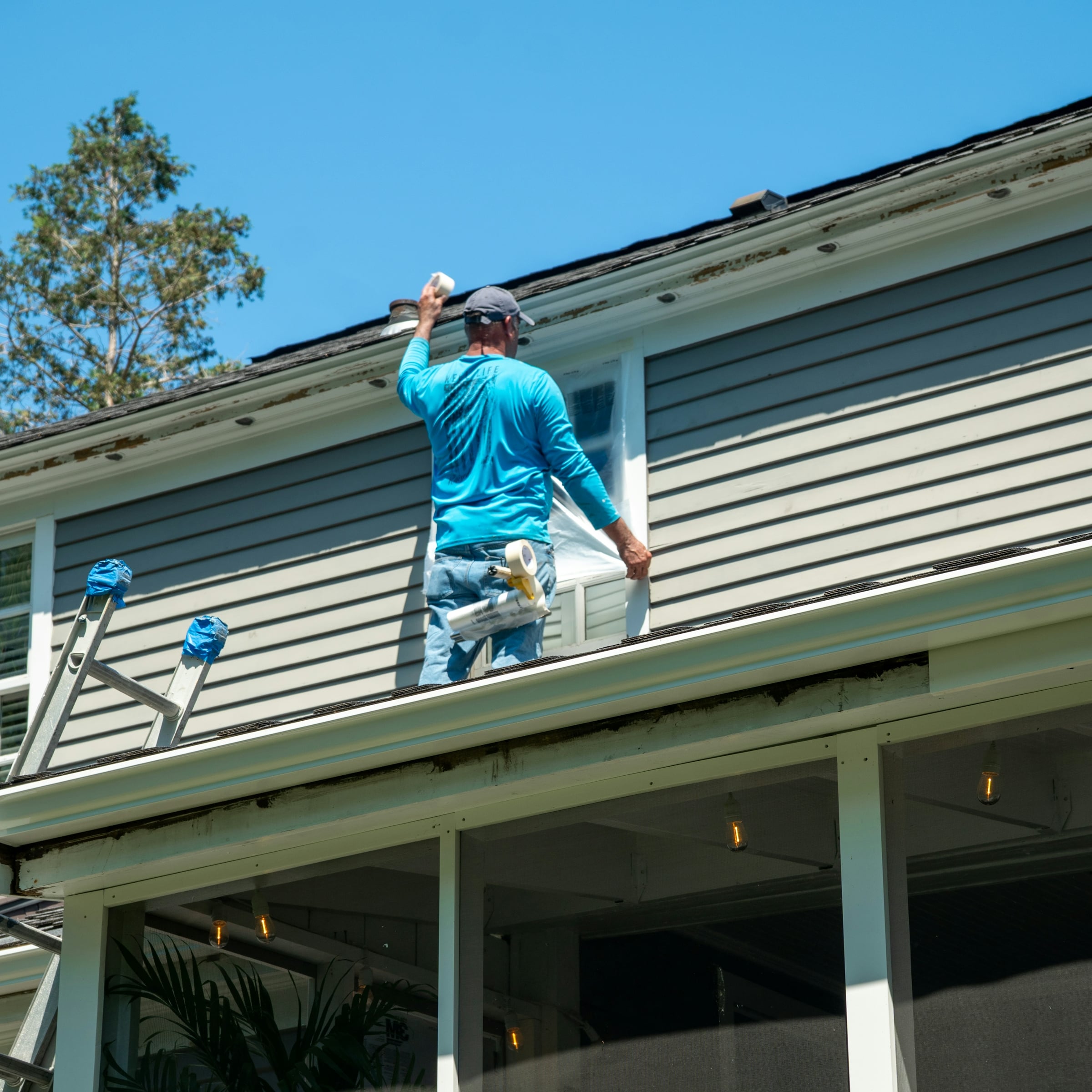A worker in blue coveralls on a ladder repairing the eaves of a house