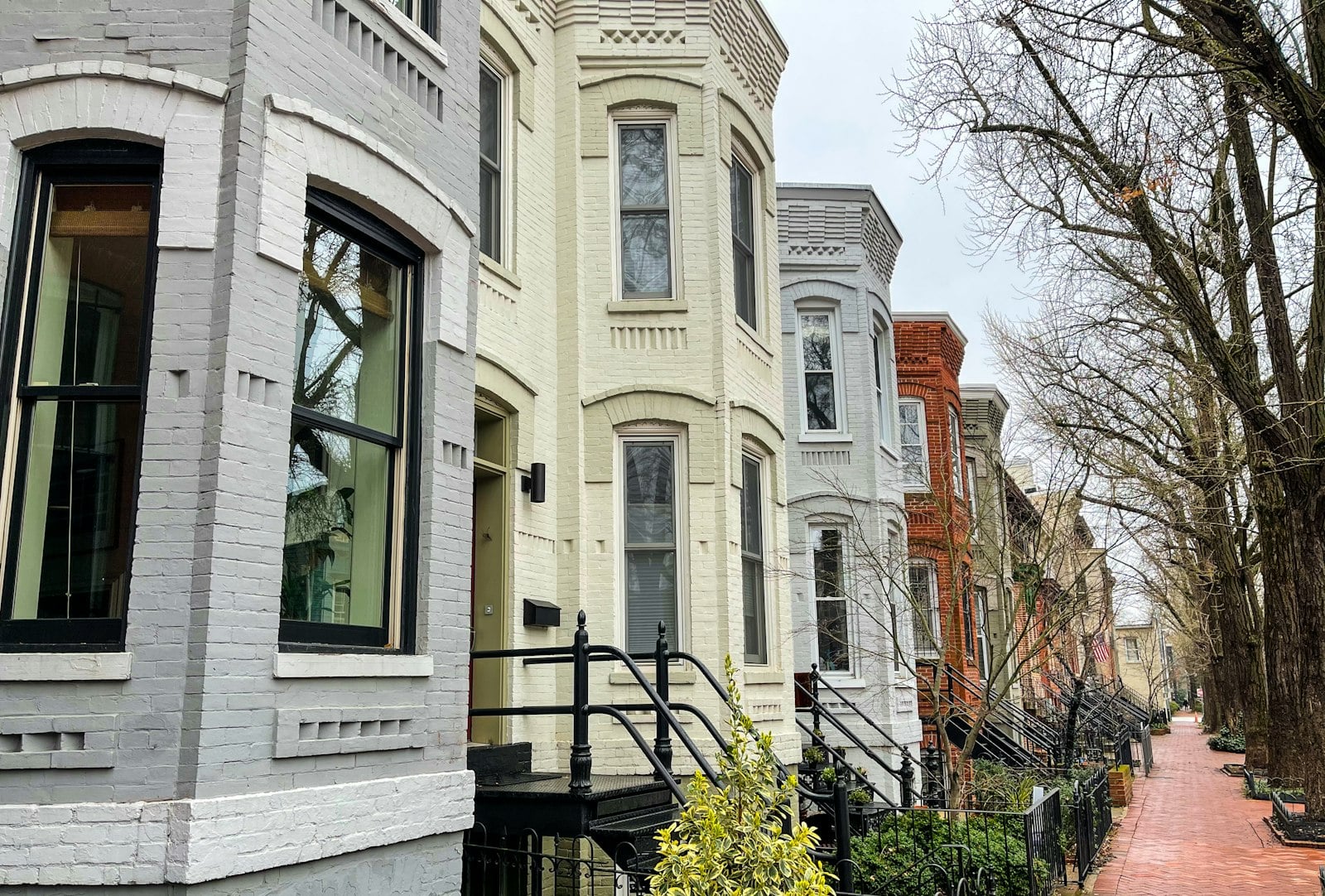Row of attached pale brick and painted townhouses with bay windows along a tree-lined brick sidewalk