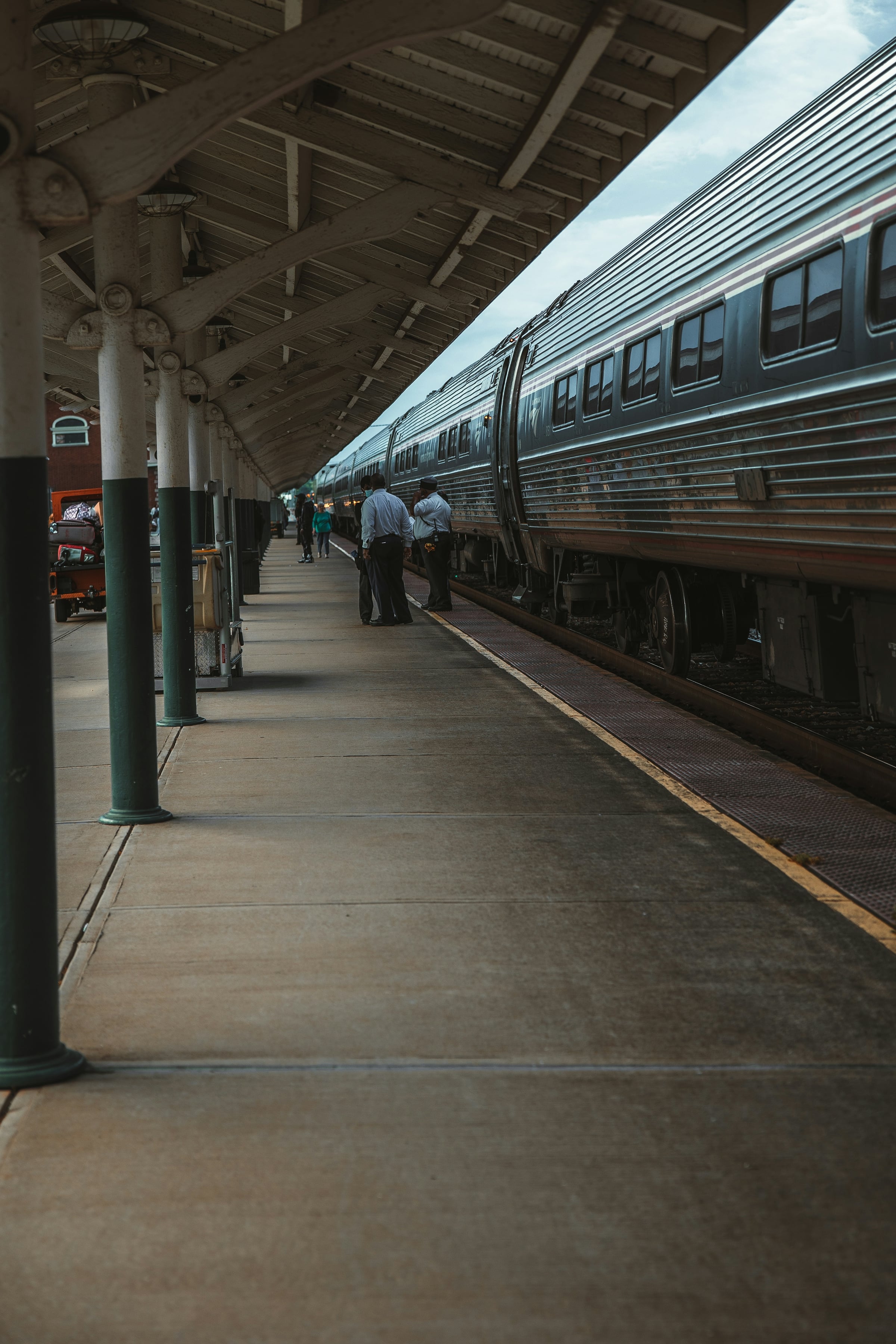 Silver passenger train stopped beside a covered station platform with green support columns, with passengers and staff standing near the cars