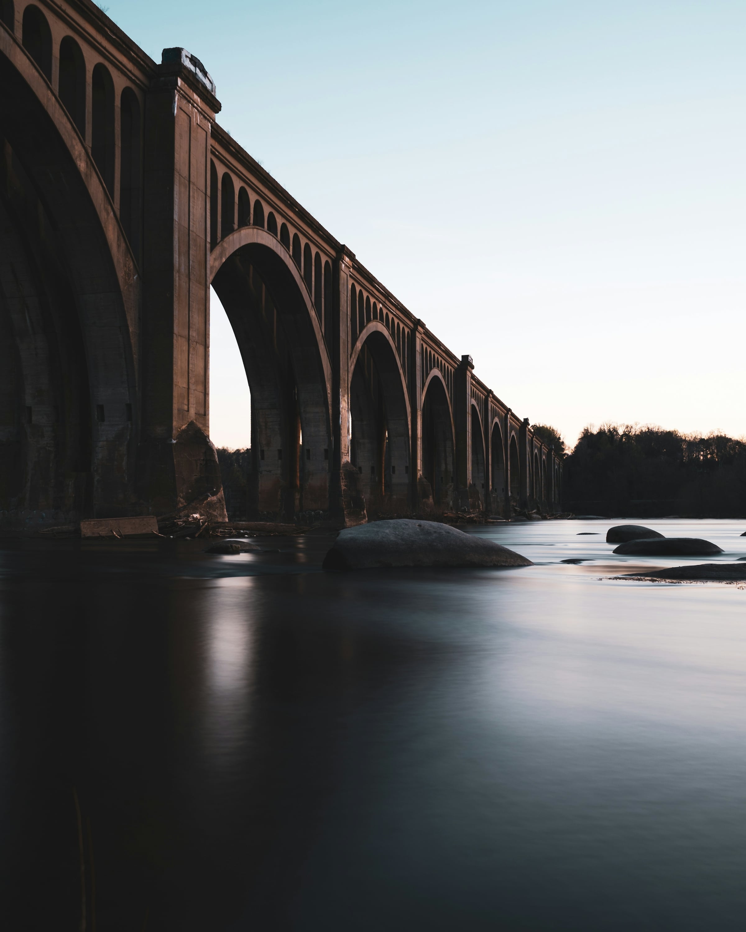 Tall brick railroad viaduct with large arches stretching across a calm river at dusk, with rocks in the water below