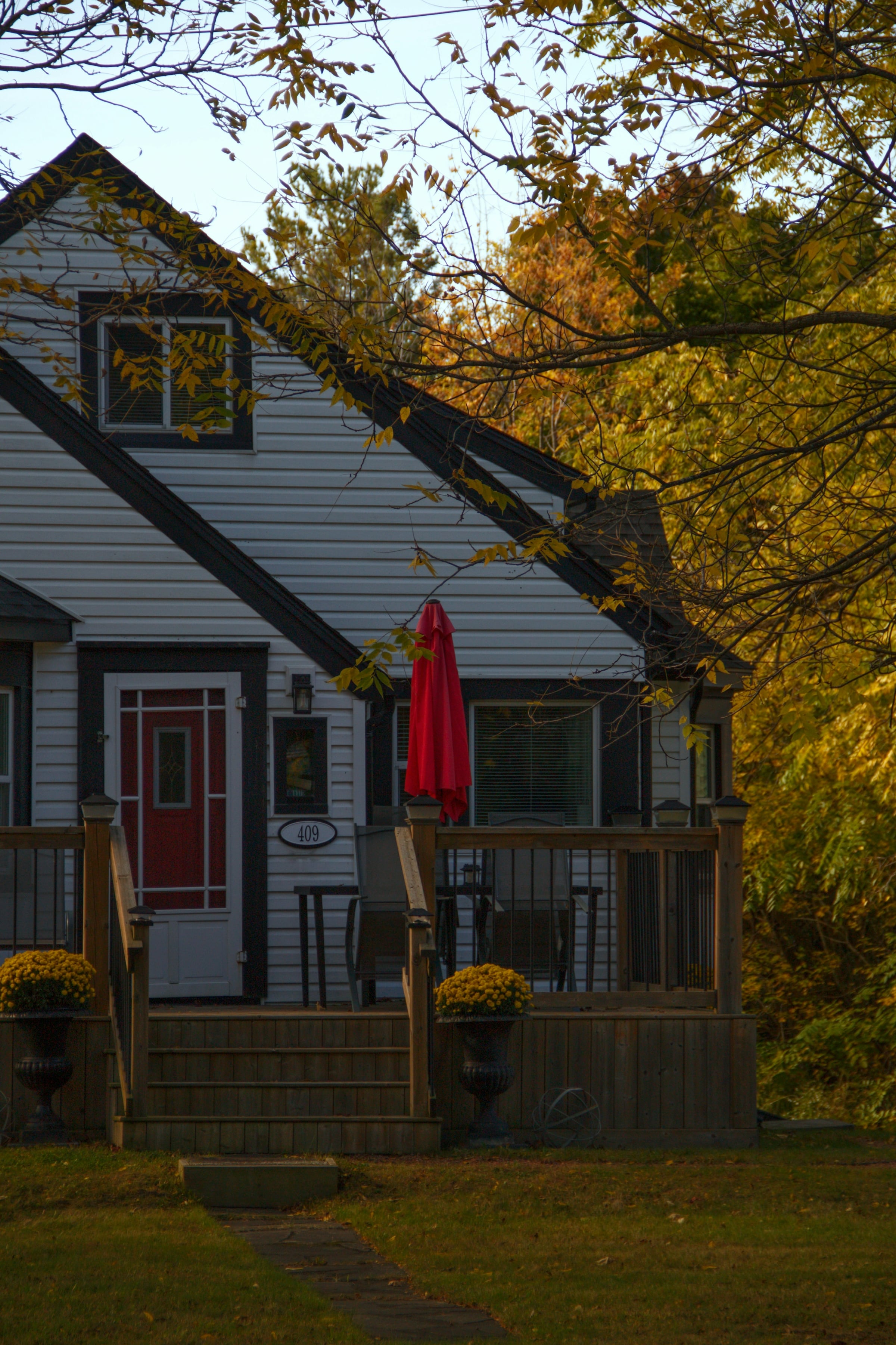 Gray-sided house with a covered front porch, wooden steps and railing, and golden autumn trees behind it