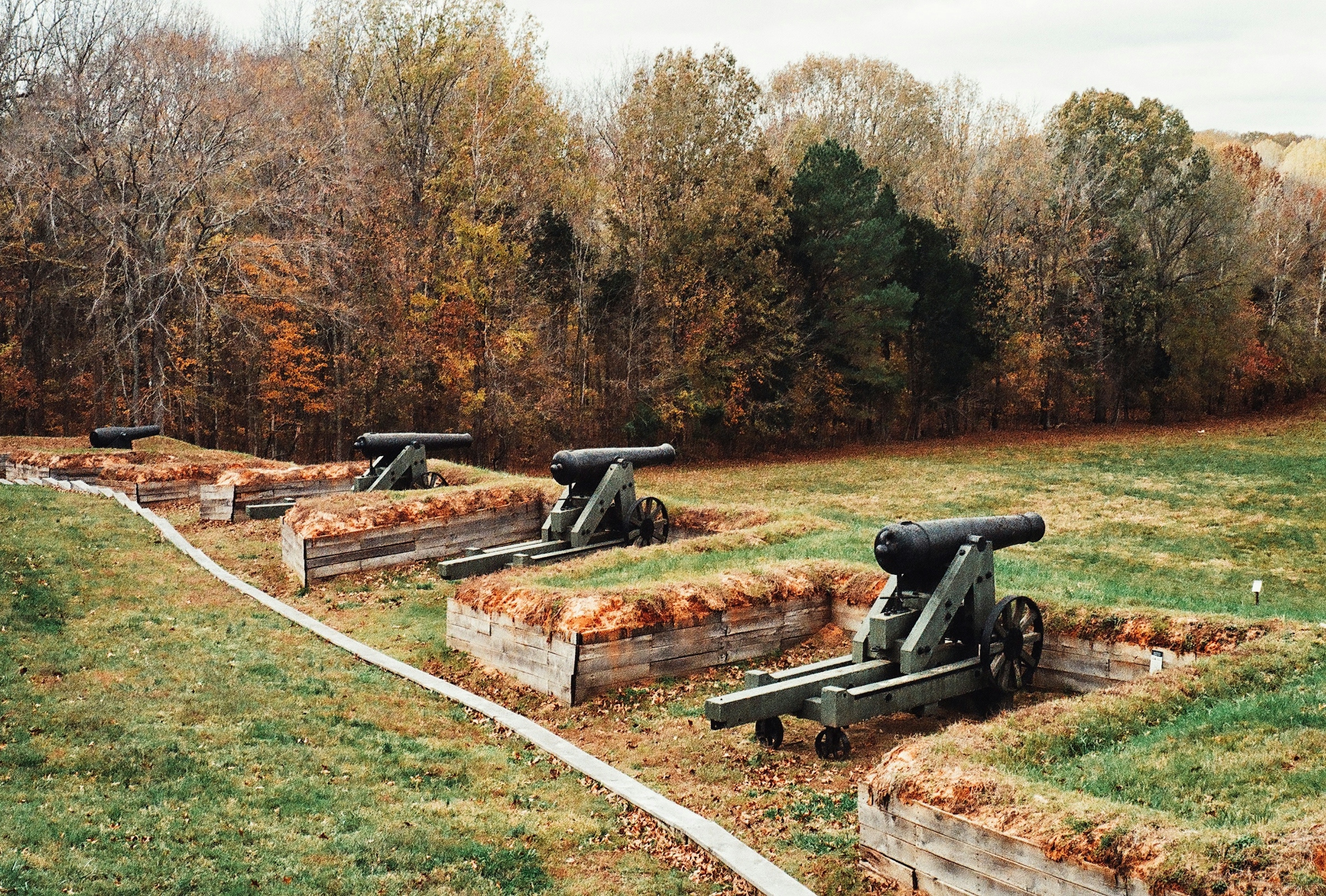 Black cannons set in earthwork gun positions beside a mowed path, with autumn-colored trees behind