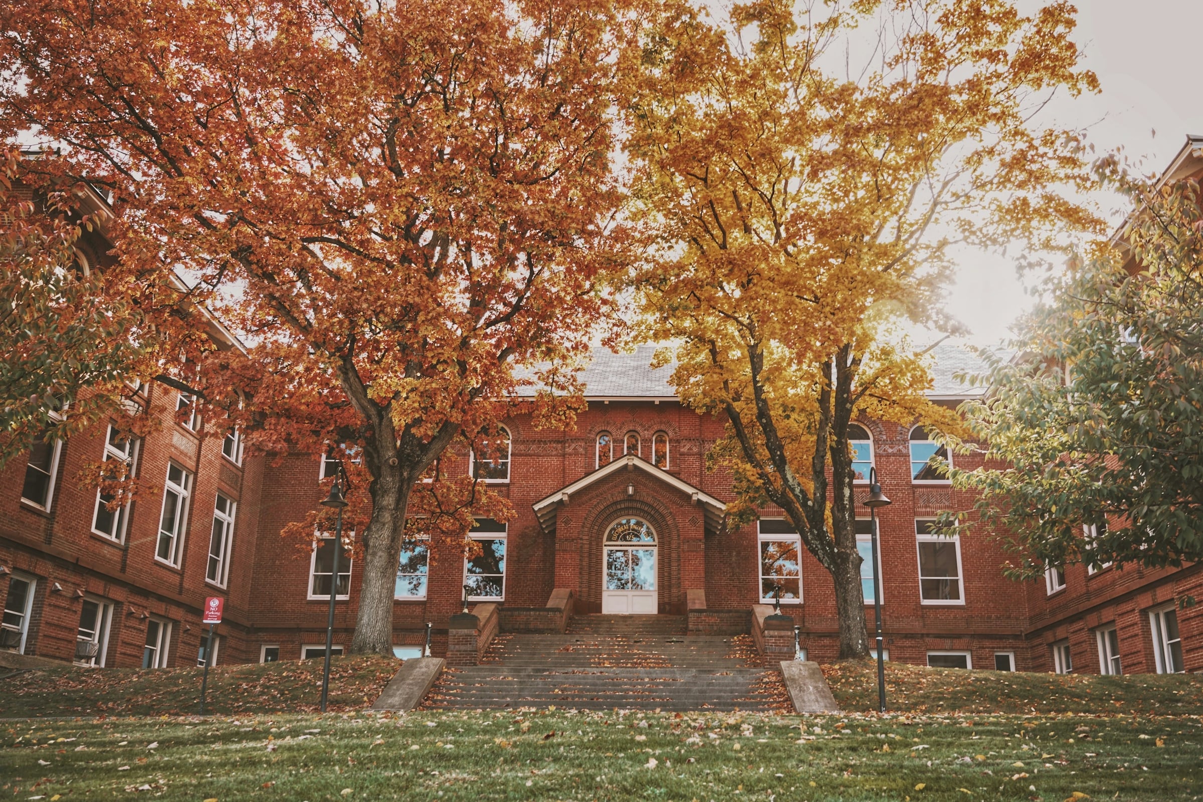 Red brick school building with an arched entrance behind trees in orange and gold autumn foliage