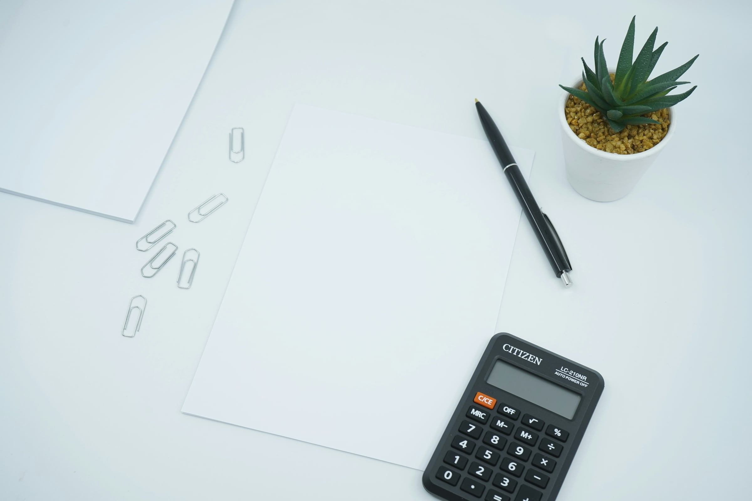 Black pocket calculator, pen and loose sheets of paper on a white desk beside a small potted succulent