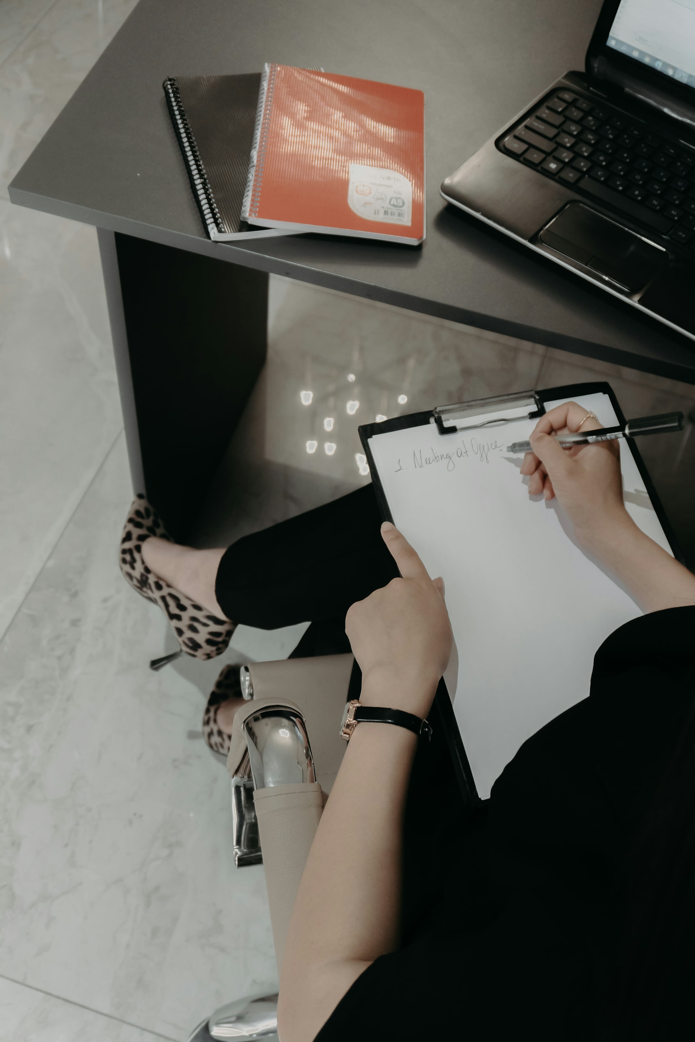Person seated at a desk writing a checklist on a clipboard with a pen, a notebook and laptop on the desk beside them