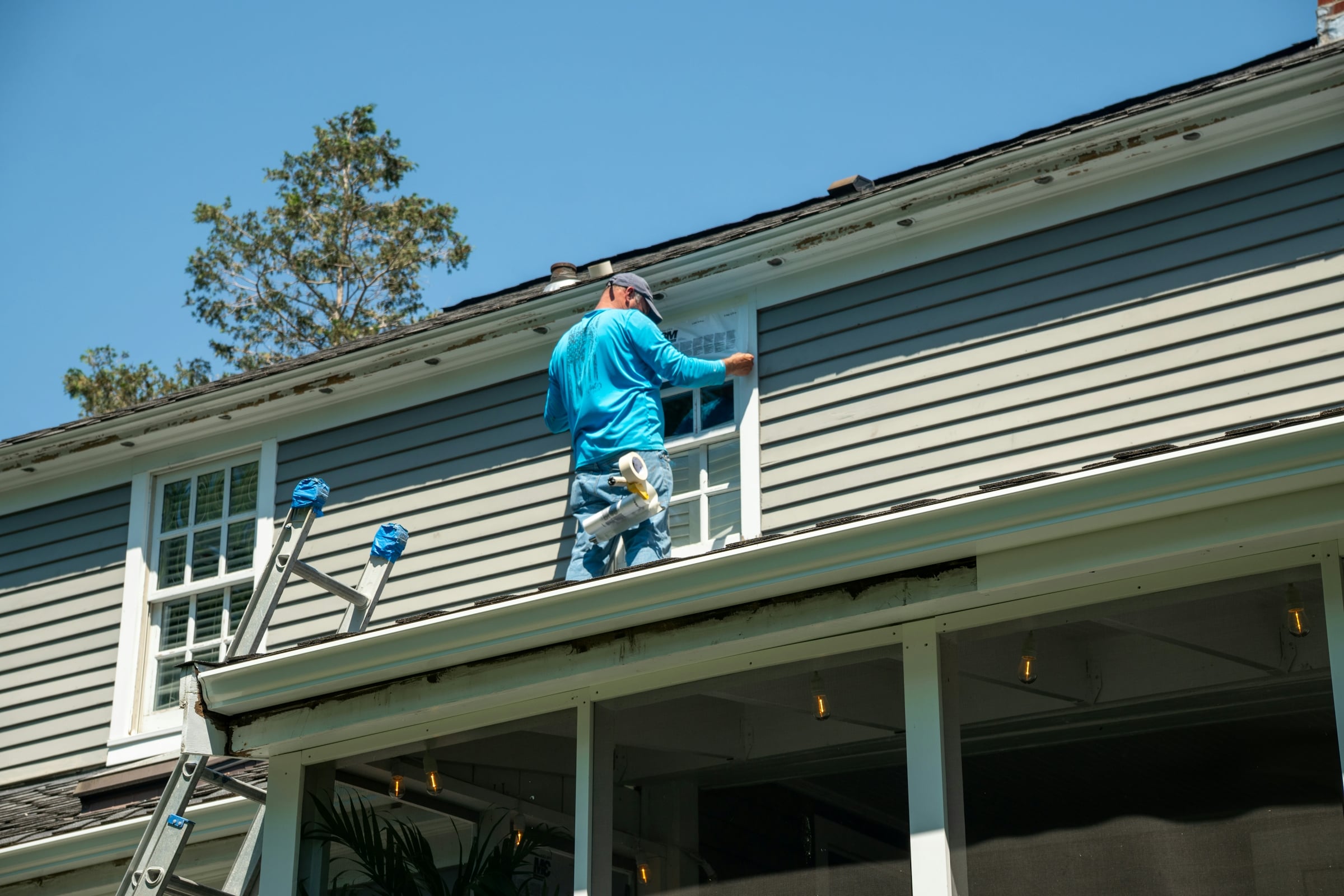 Contractor in a blue shirt working on the window trim of a gray-sided house from a ladder, with the gutter and porch roof below him