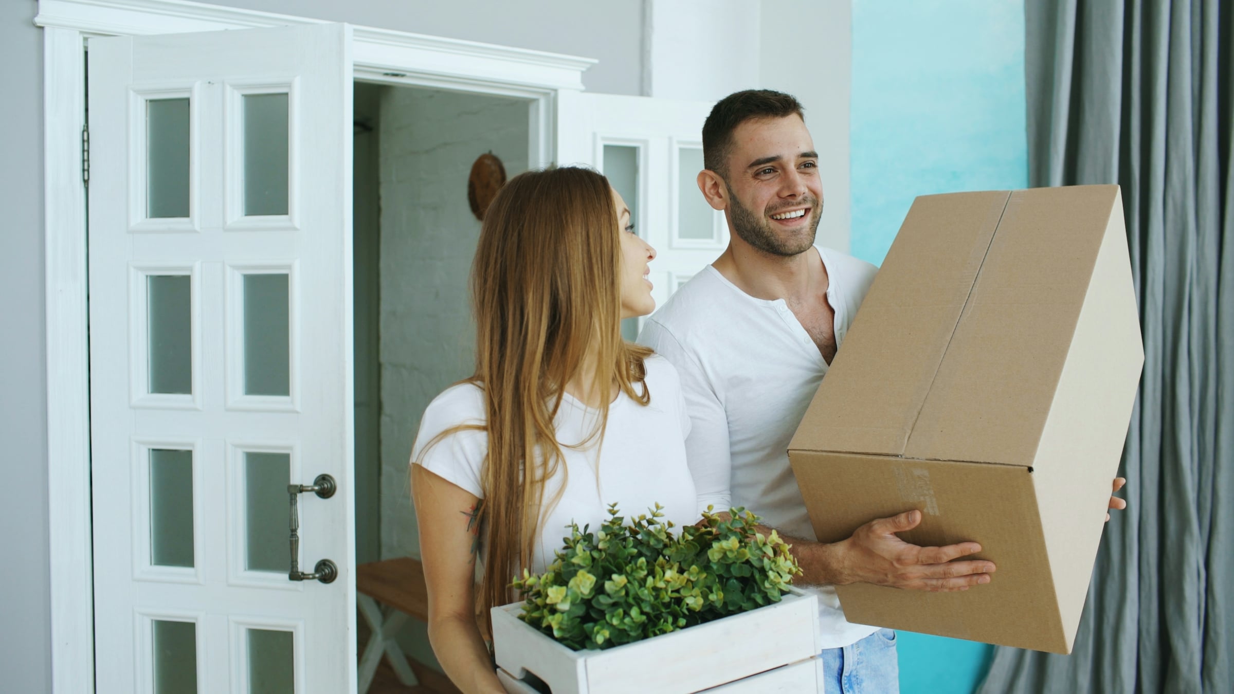 Couple carrying a cardboard moving box and a potted plant through the doorway of a new home