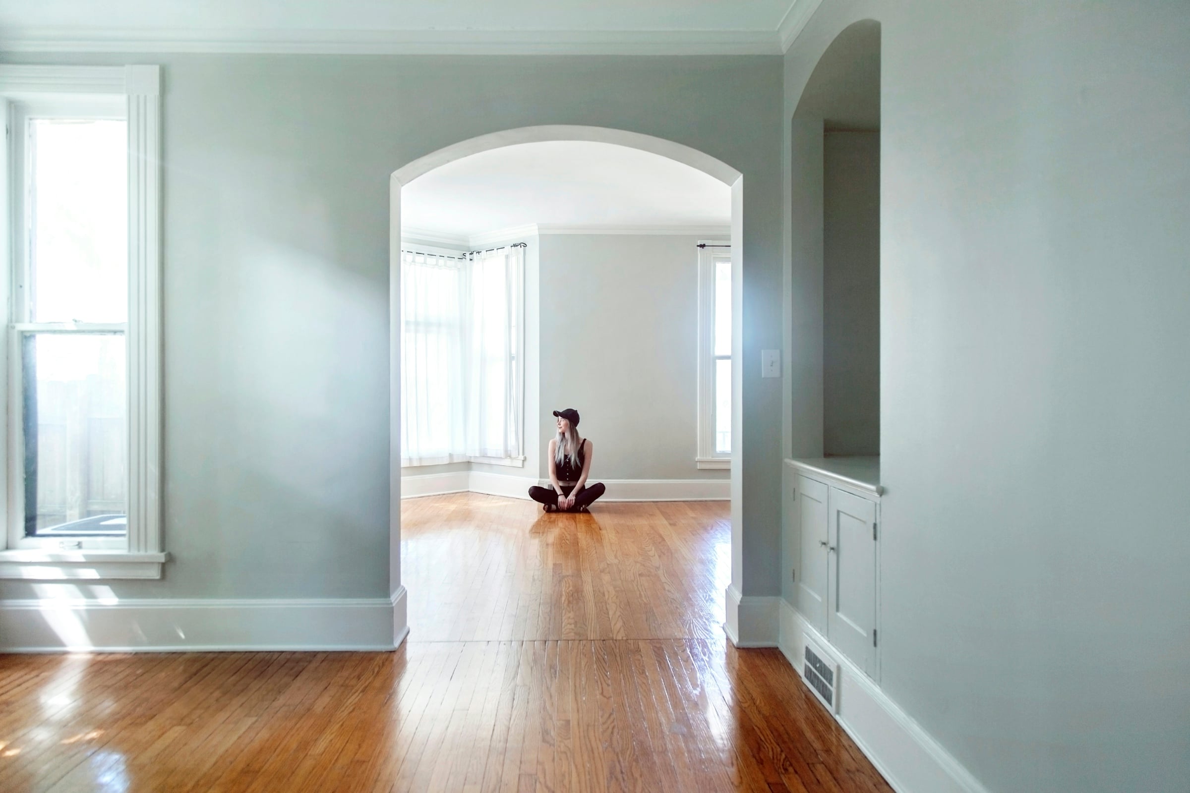 Empty hardwood-floored rooms with a wide arched doorway, built-in cabinet and tall windows in a vacant house