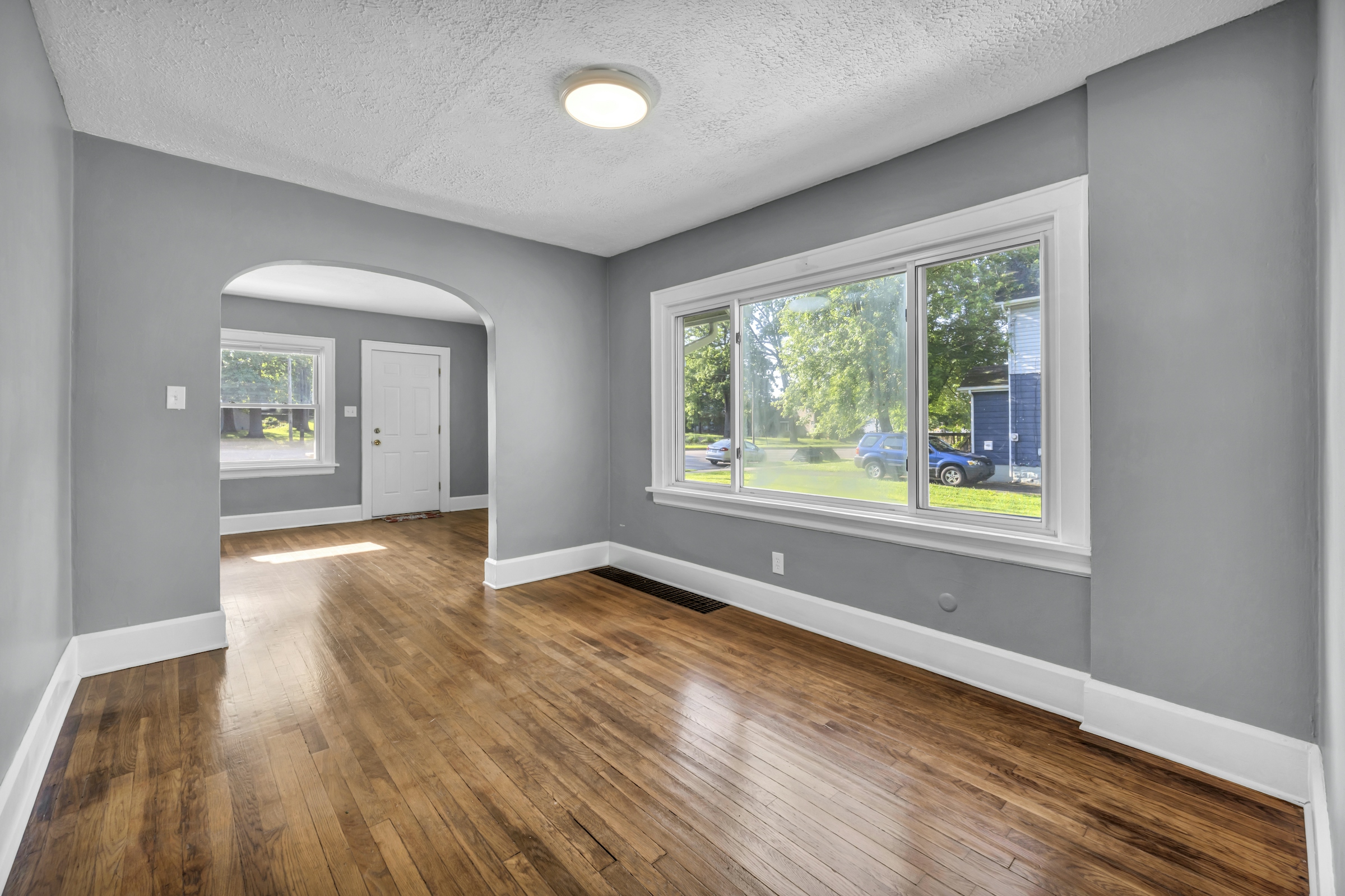 Empty living room with gray walls, white trim, a large front window and refinished hardwood floors