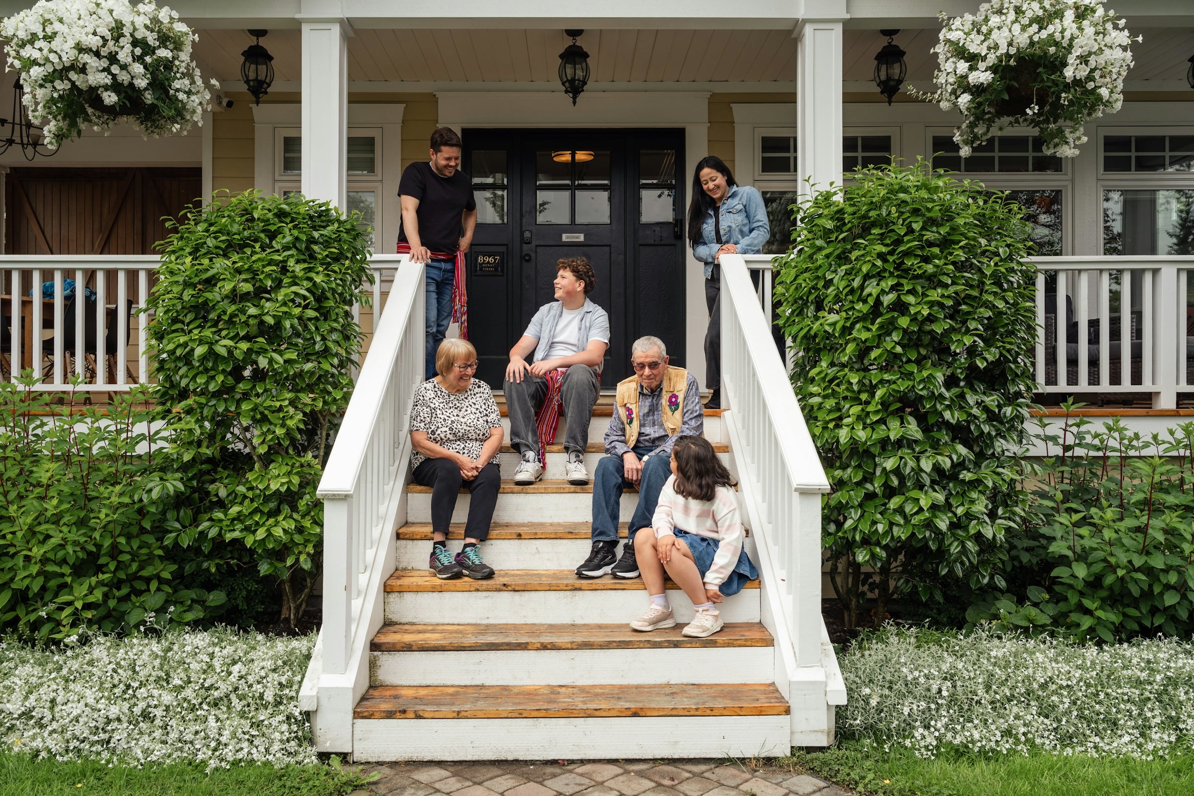 Several generations of a family sitting and standing on the white front steps of a porch with hanging flower baskets