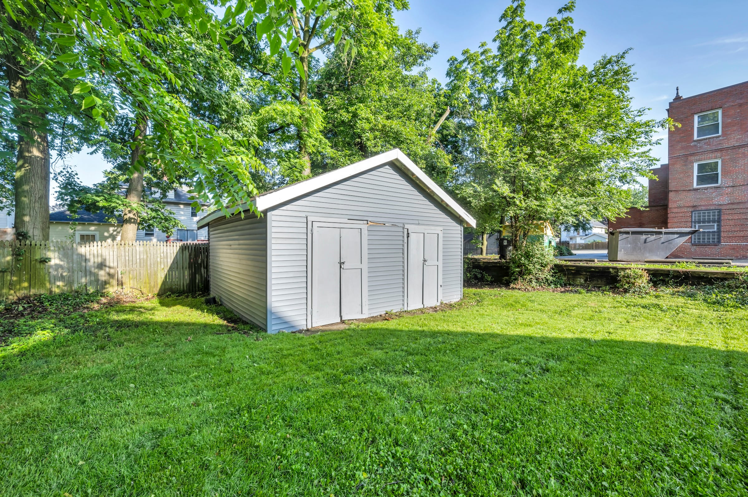 Gray vinyl-sided detached shed with double doors on a mowed lawn beneath mature shade trees