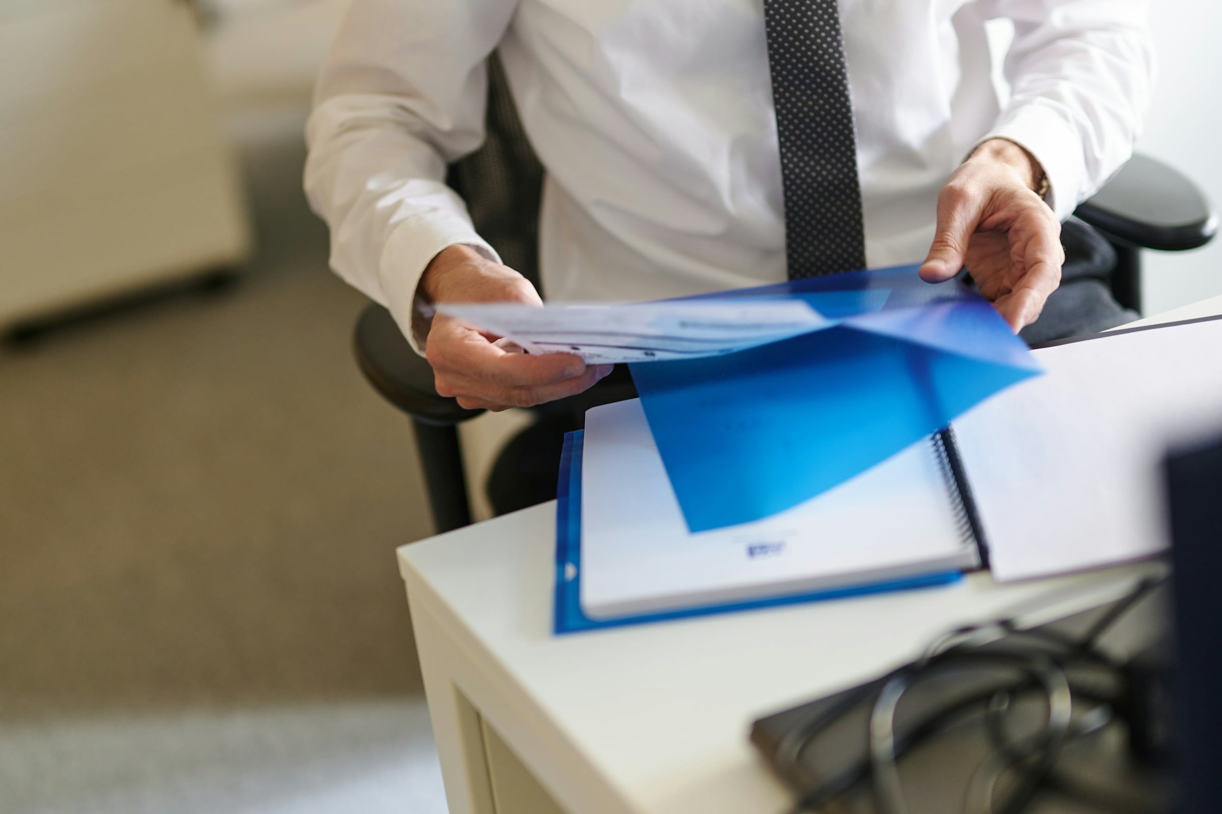 A person in a white shirt and tie sorting papers from a blue folder at a desk
