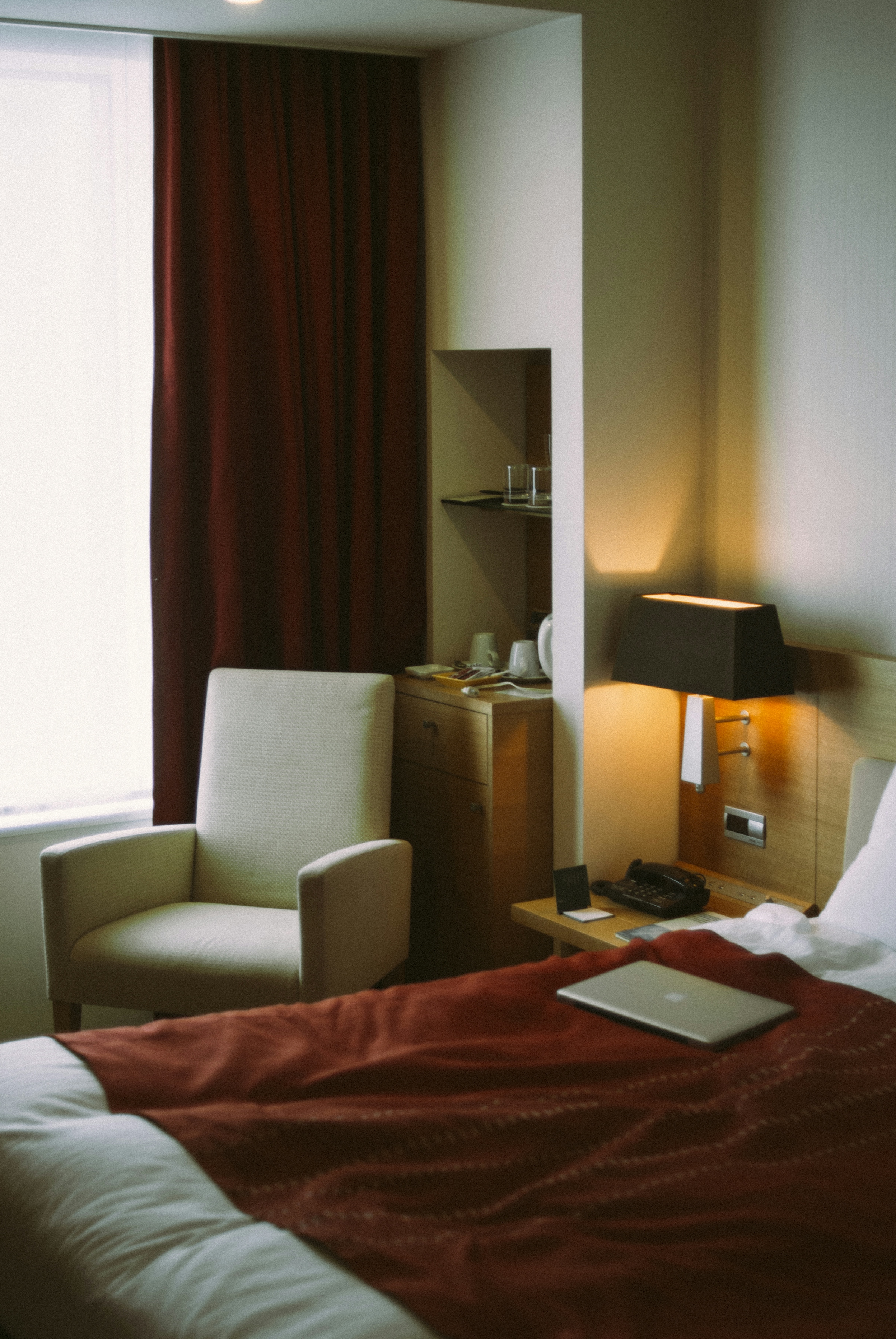 Hotel room with a cream armchair beside a curtained window and a lit bedside lamp