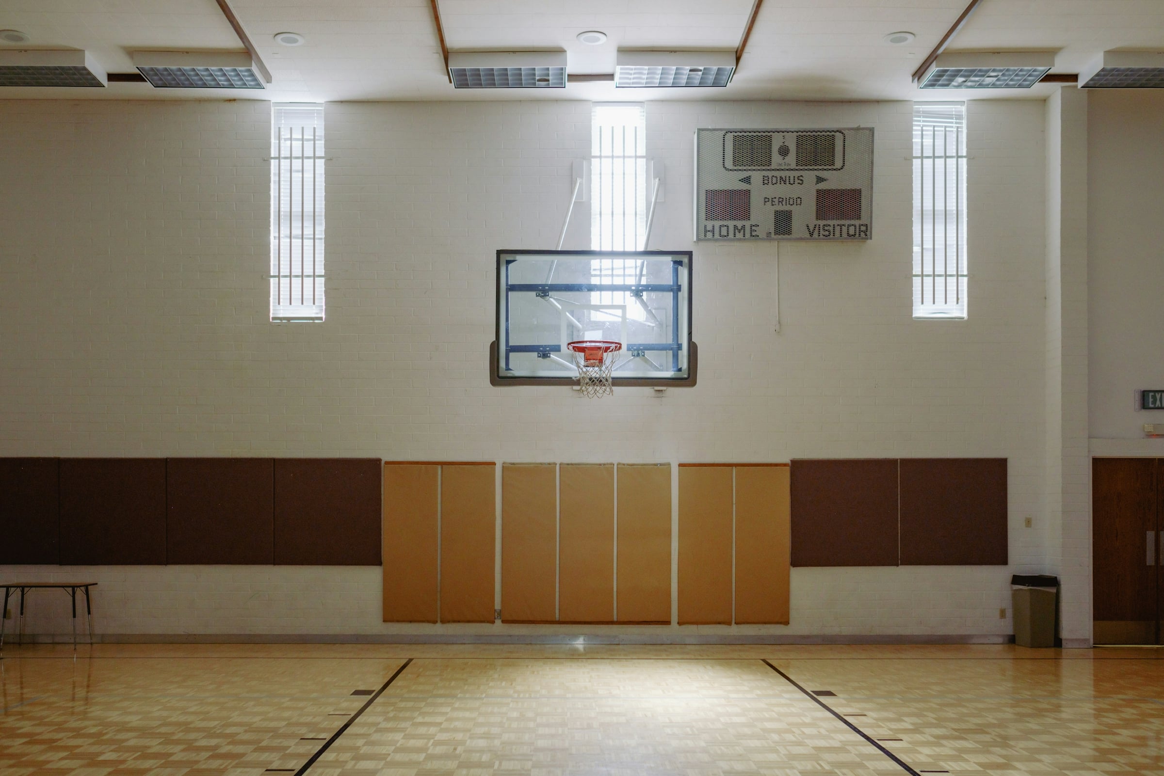 An empty indoor gymnasium with a wood floor, a basketball hoop and a scoreboard on the wall