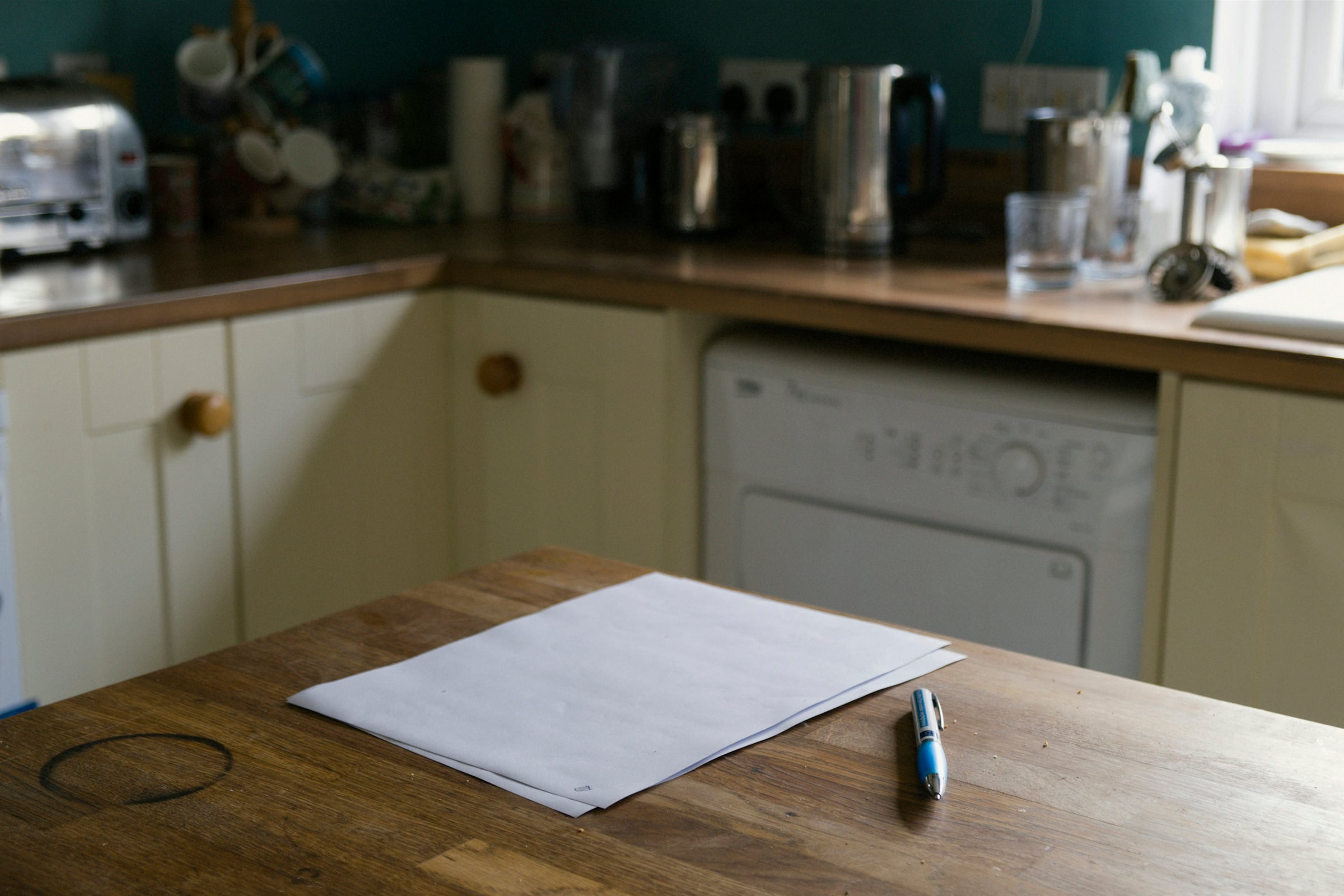 Printed pages and a pen resting on a wooden kitchen table, with a cream-colored kitchen counter and dishwasher behind them