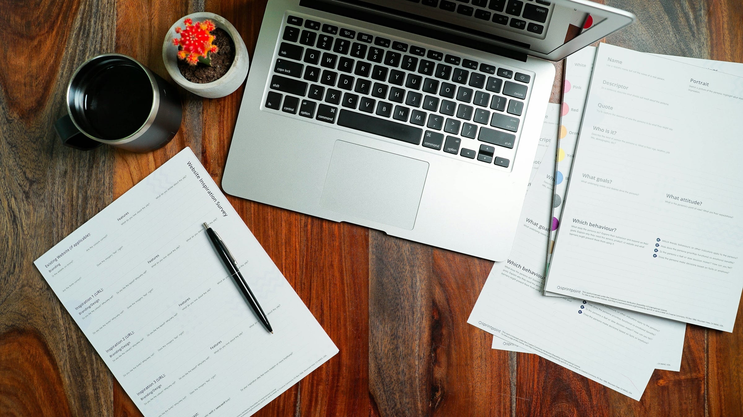 Open laptop on a wooden desk beside printed paperwork, a pen and a coffee cup