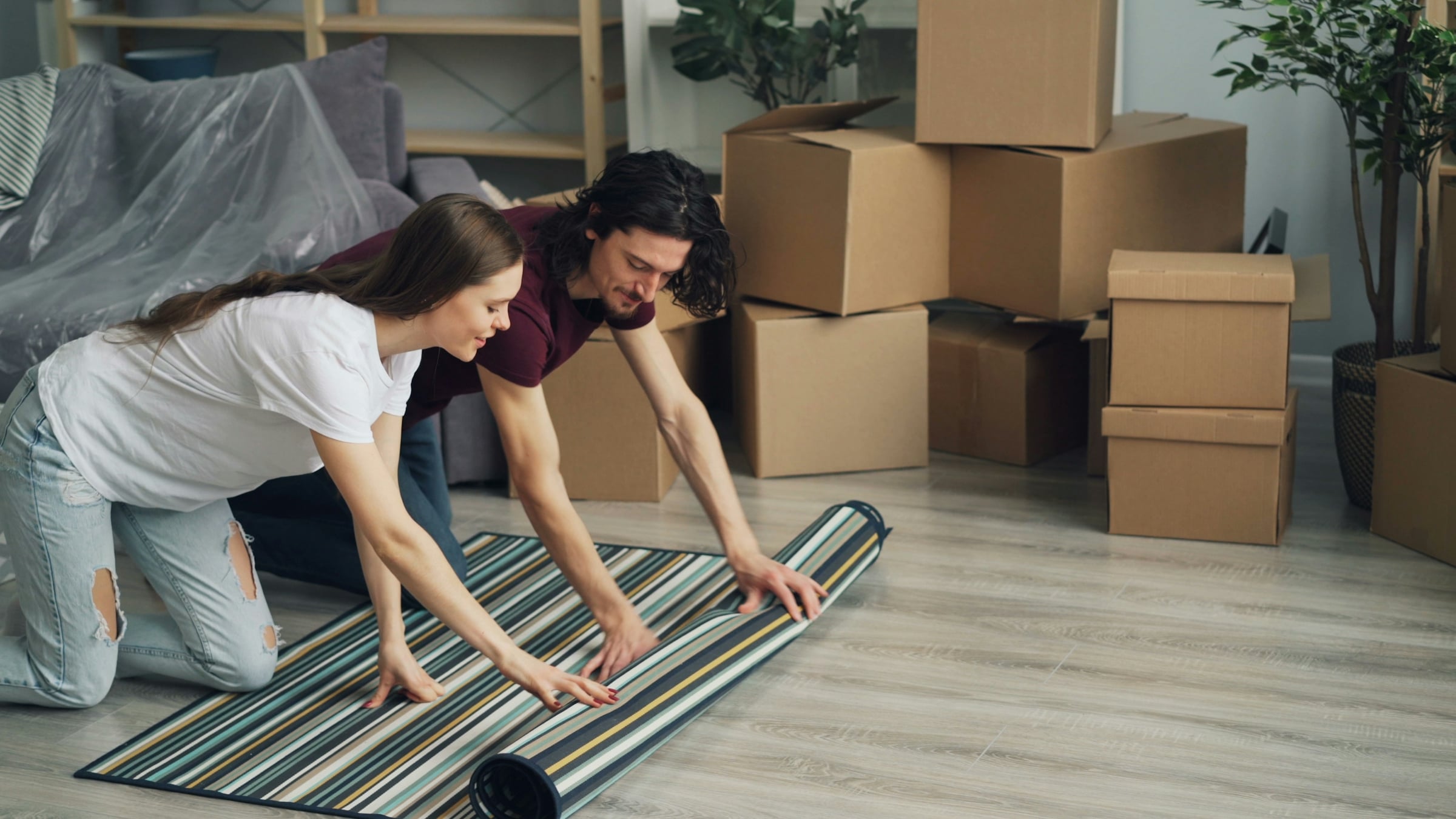 Two people kneeling on a pale wood floor unrolling a striped rug beside a stack of cardboard moving boxes in a living room