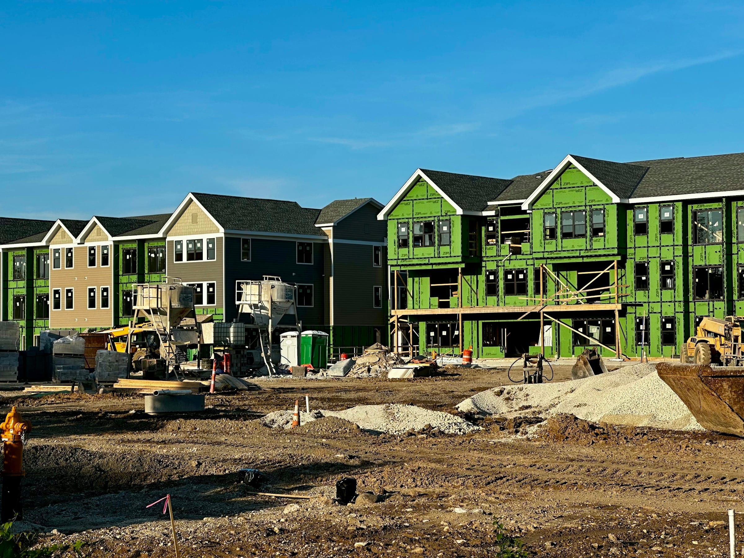 Rows of new townhomes under construction, wrapped in green sheathing, with an excavator and bare soil in front