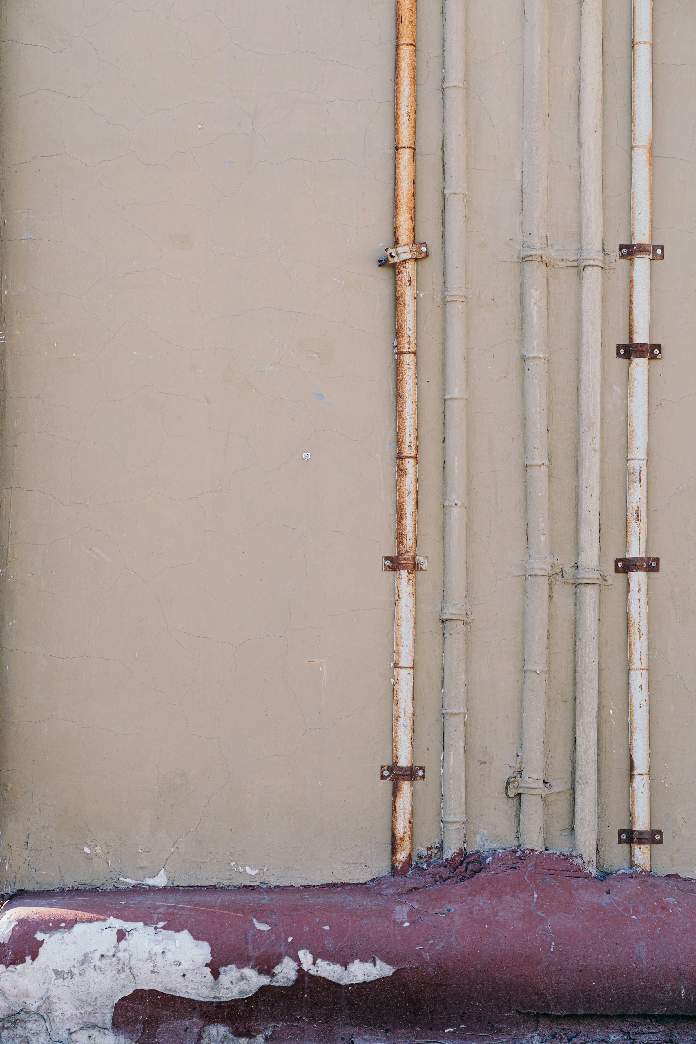 Old painted pipes running down a beige wall, some of them rusted, held by metal brackets