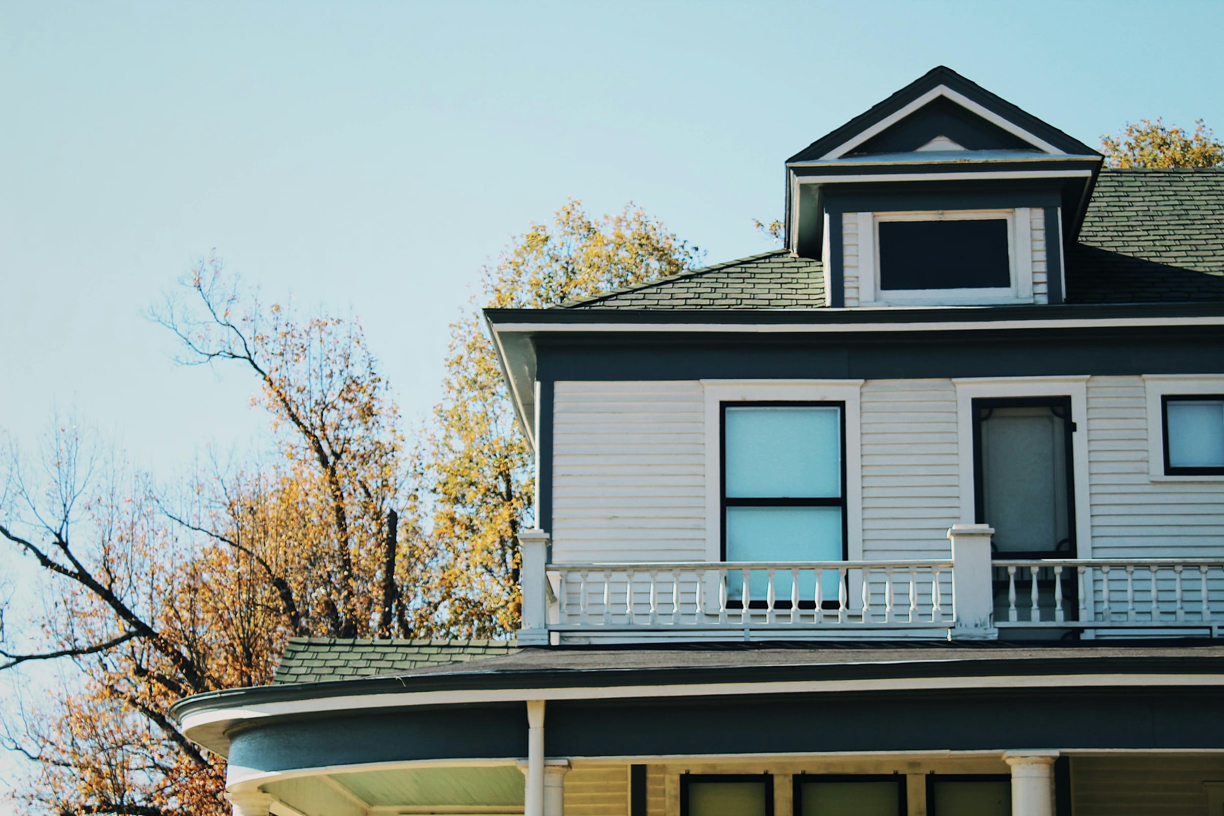 Two-story white frame house with a wraparound porch and dormer window under a blue sky, with autumn trees behind
