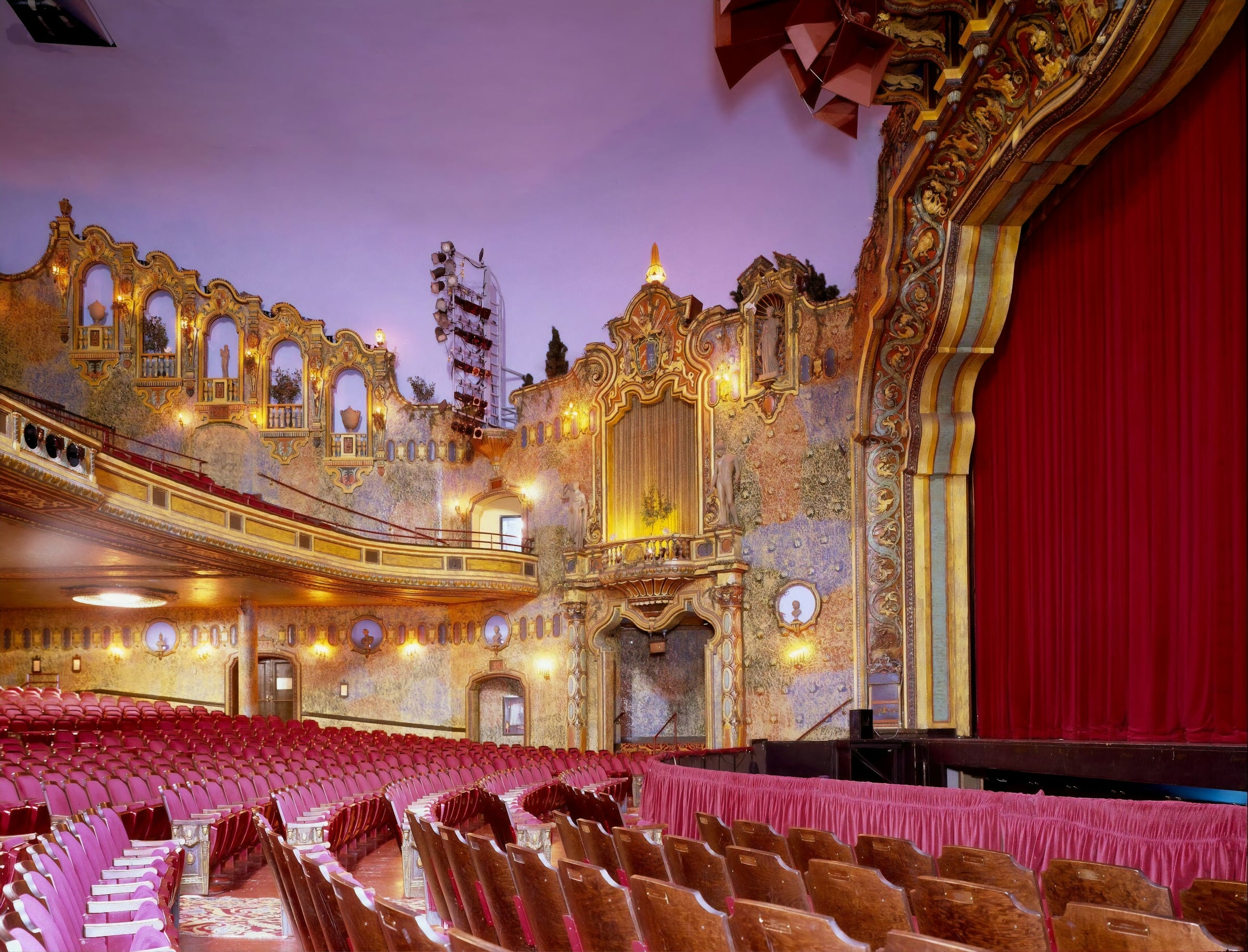 Ornate historic theater auditorium with a gilded proscenium, a red stage curtain and rows of red seats